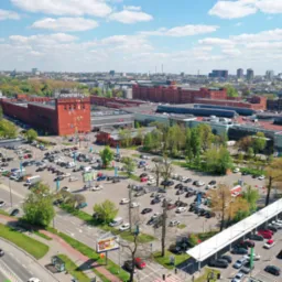 Panorama Łodzi z lotu ptaka, widok na budynek Manufaktury i parking, w tle zabudowa miejska i błękitne niebo z chmurami.
