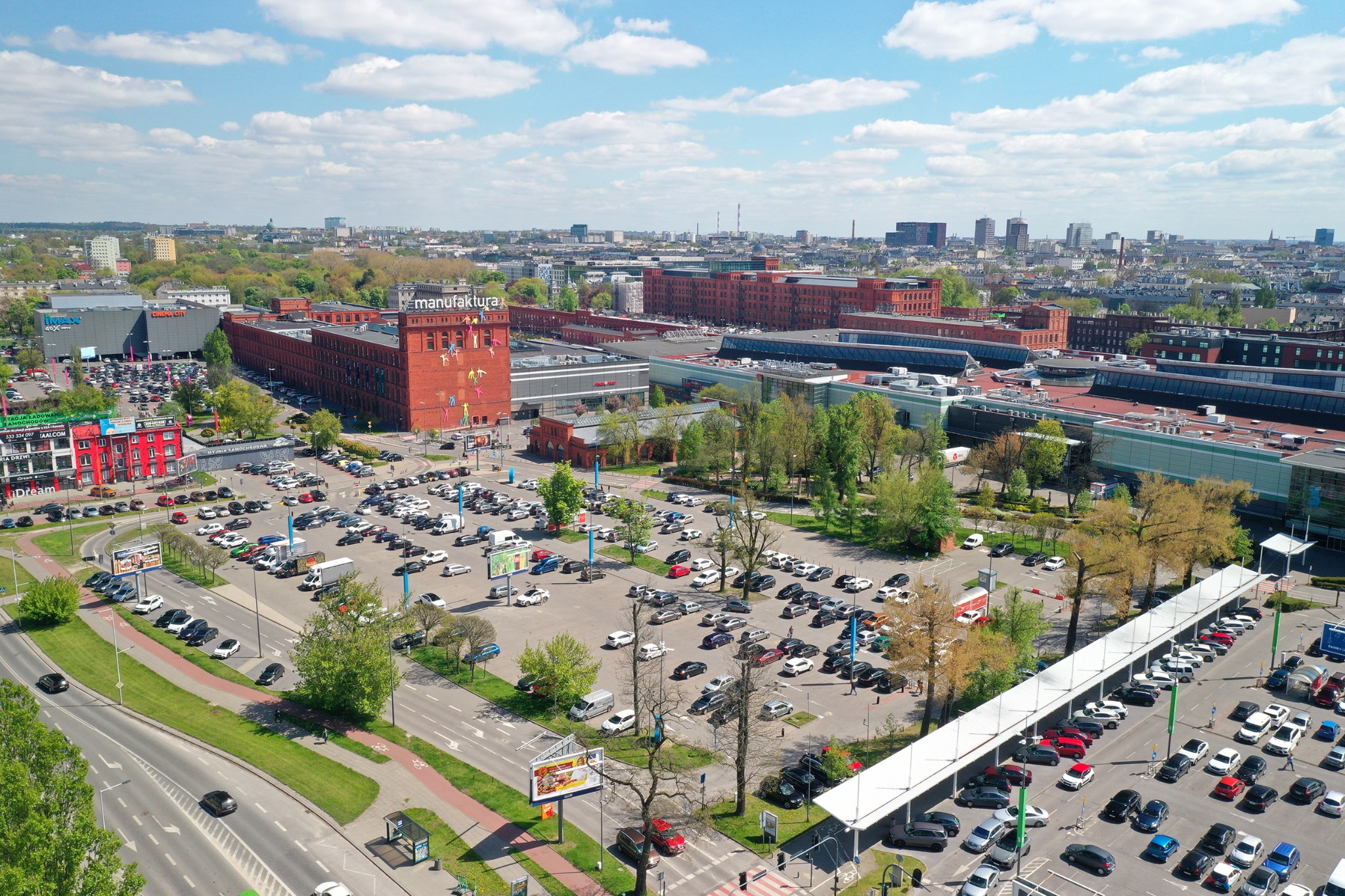 Panorama Łodzi z lotu ptaka, widok na budynek Manufaktury i parking, w tle zabudowa miejska i błękitne niebo z chmurami.