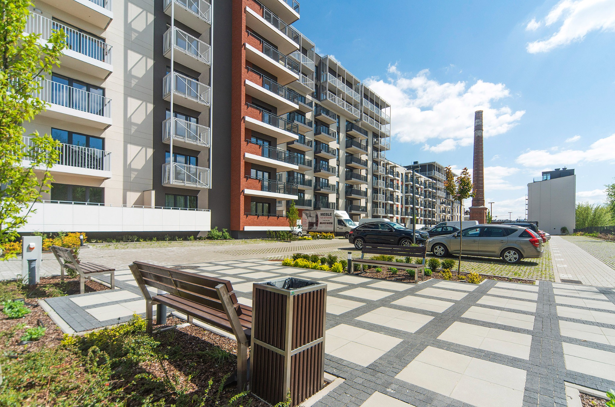 Nowoczesny budynek wielorodzinny z balkonami, widok z poziomu dziedzińca z ławkami i kostką brukową, w tle komin starej fabryki.