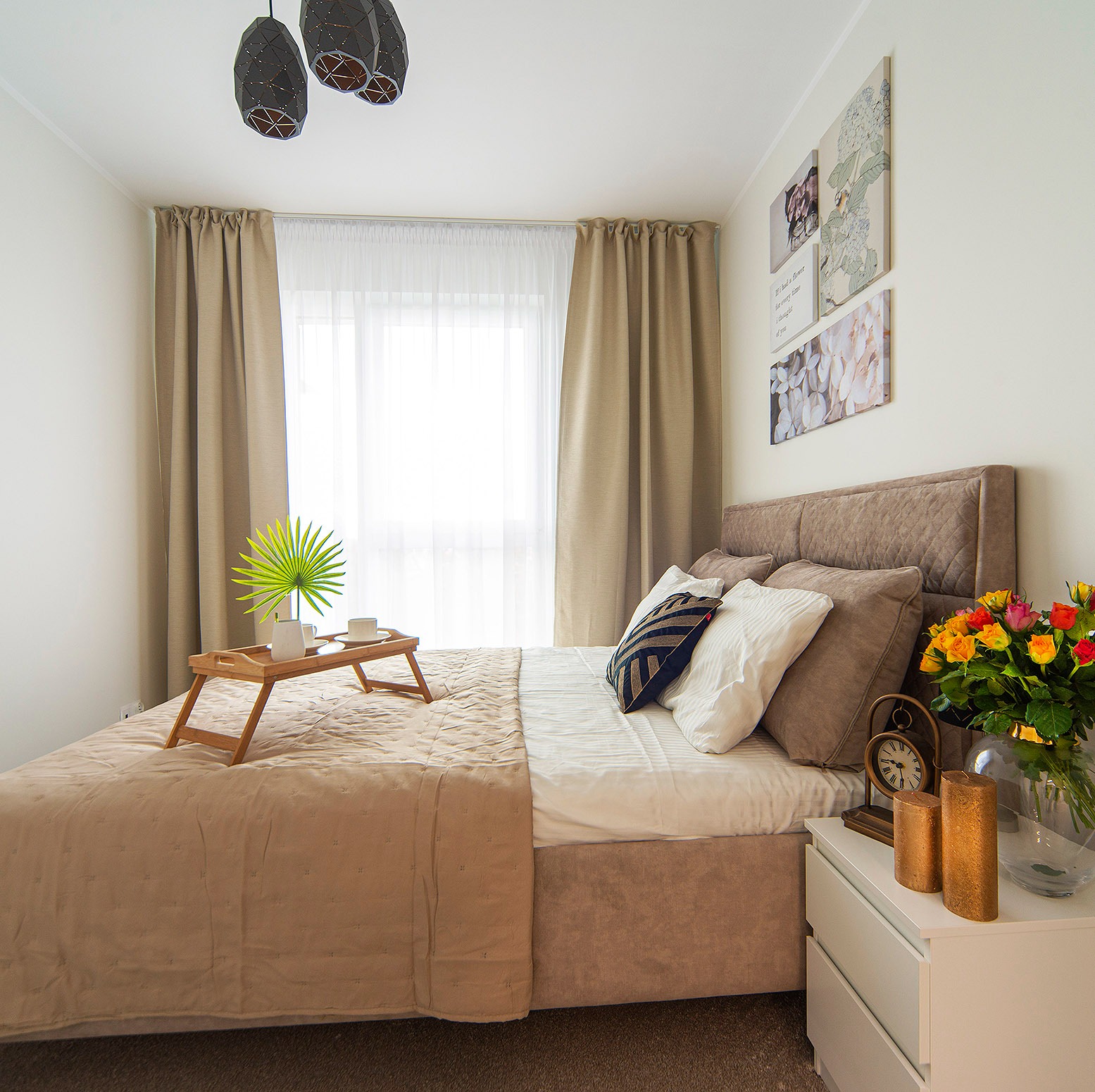 Jasna sypialnia z beżowym łóżkiem, drewnianą tacą z napojami i palmą, bukietem róż na białej szafce nocnej oraz designerskimi lampami wiszącymi