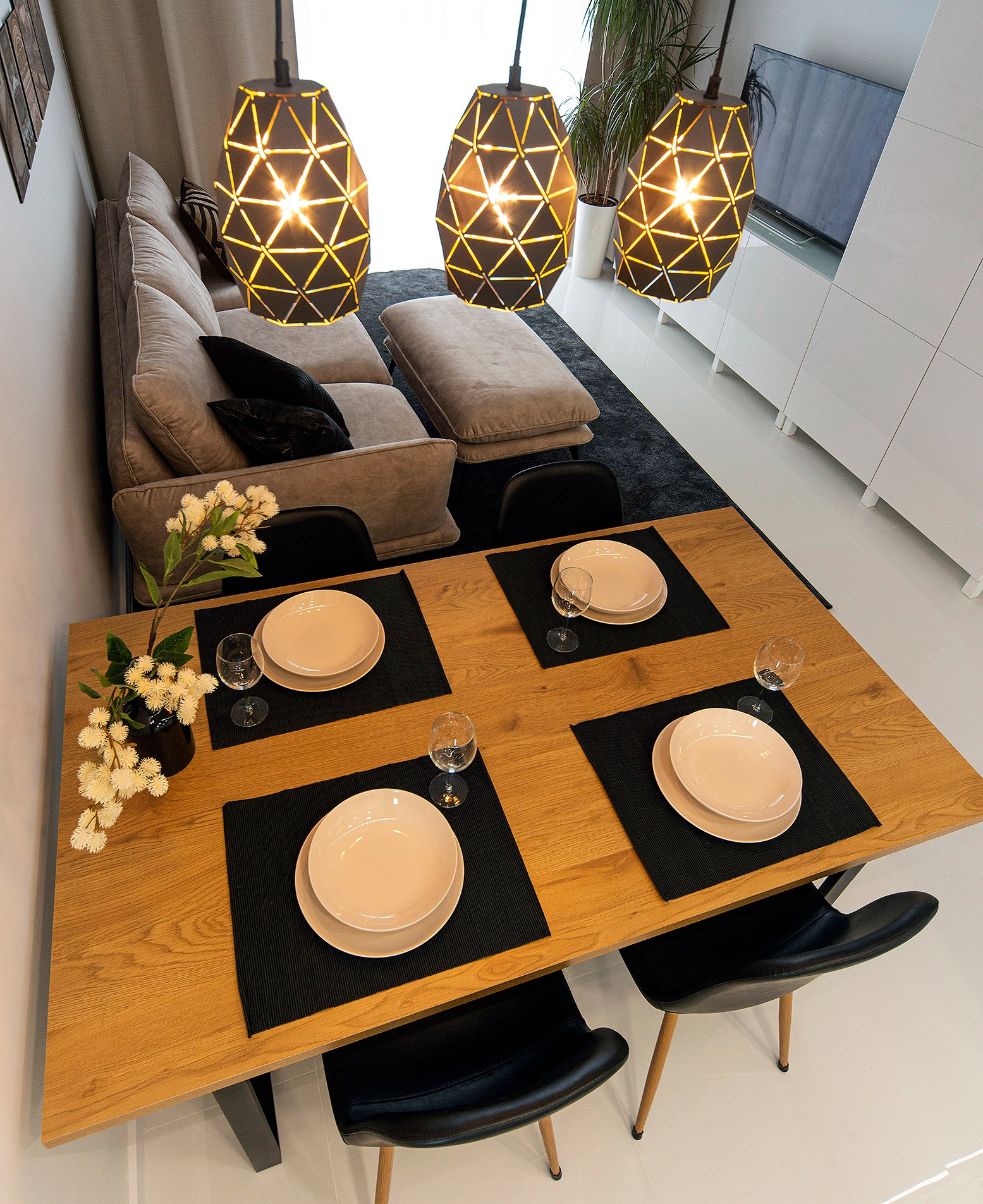 Jadalnia w nowoczesnym stylu: drewniany stół z czterema nakryciami, czarne podkładki, jasne talerze i kieliszki, designerskie lampy wiszące, szara sofa i białe meble w tle.