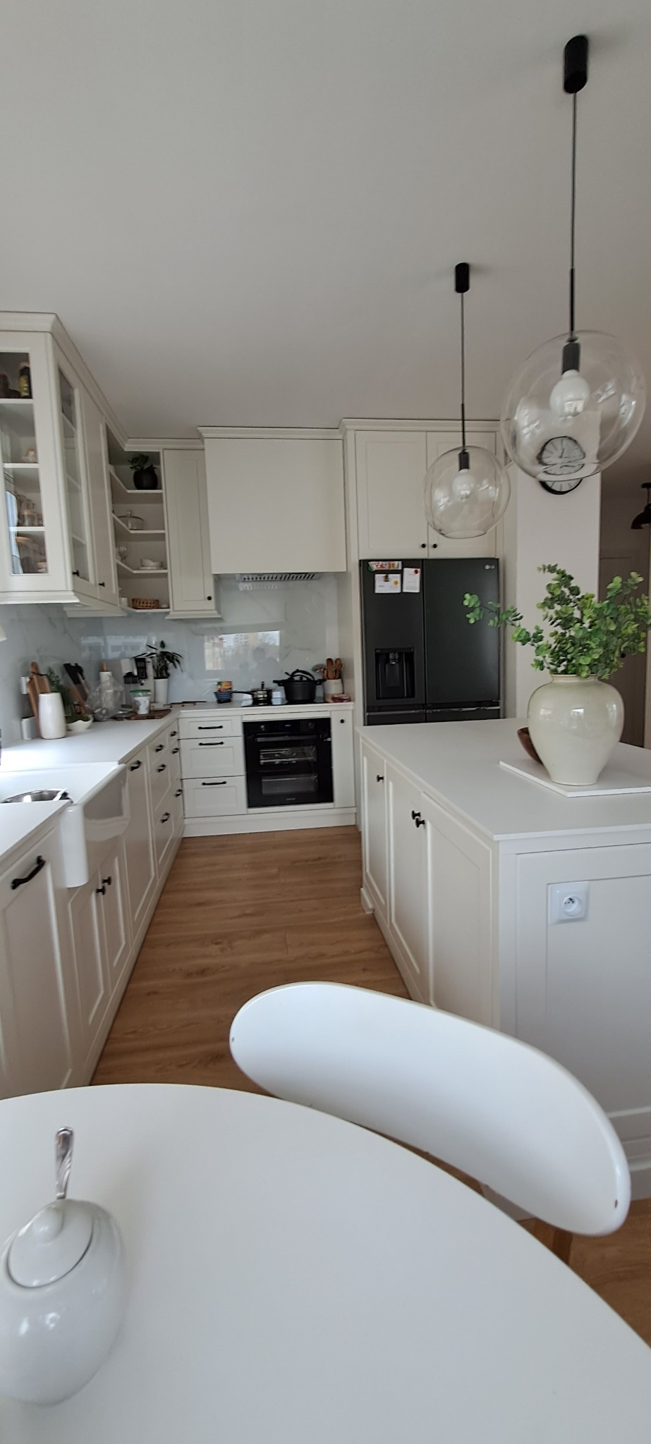 Jasna, przestronna kuchnia z białymi szafkami, drewnianym blatem i wyspą kuchenną, oświetlona wiszącymi lampami w kształcie kul, z widocznym fragmentem białego stołu i krzesła.