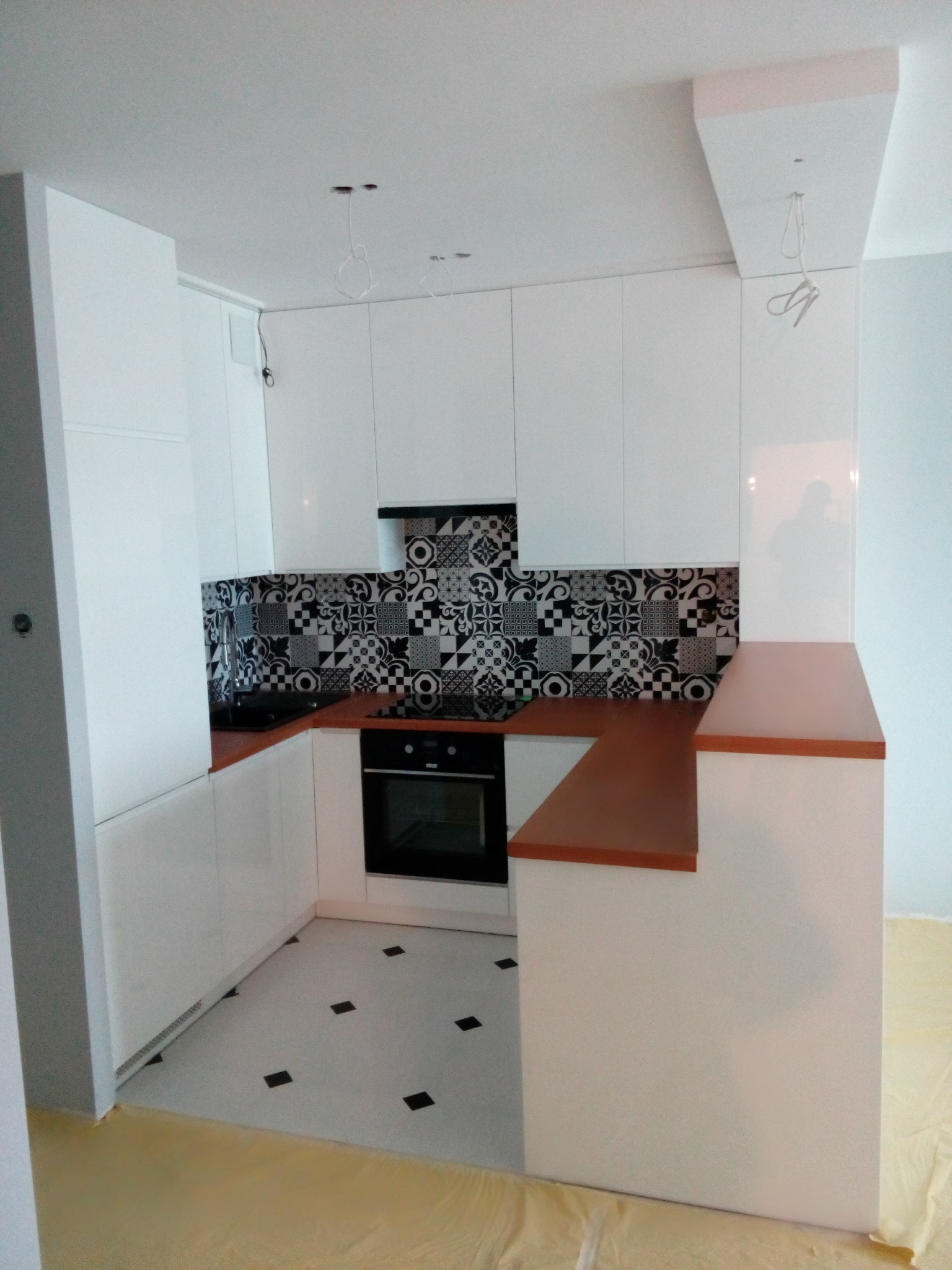 Nowoczesna biała kuchnia z brązowym blatem i wzorzystą mozaiką nad blatem, widoczne niedokończone podłączenia oświetlenia sufitowego.