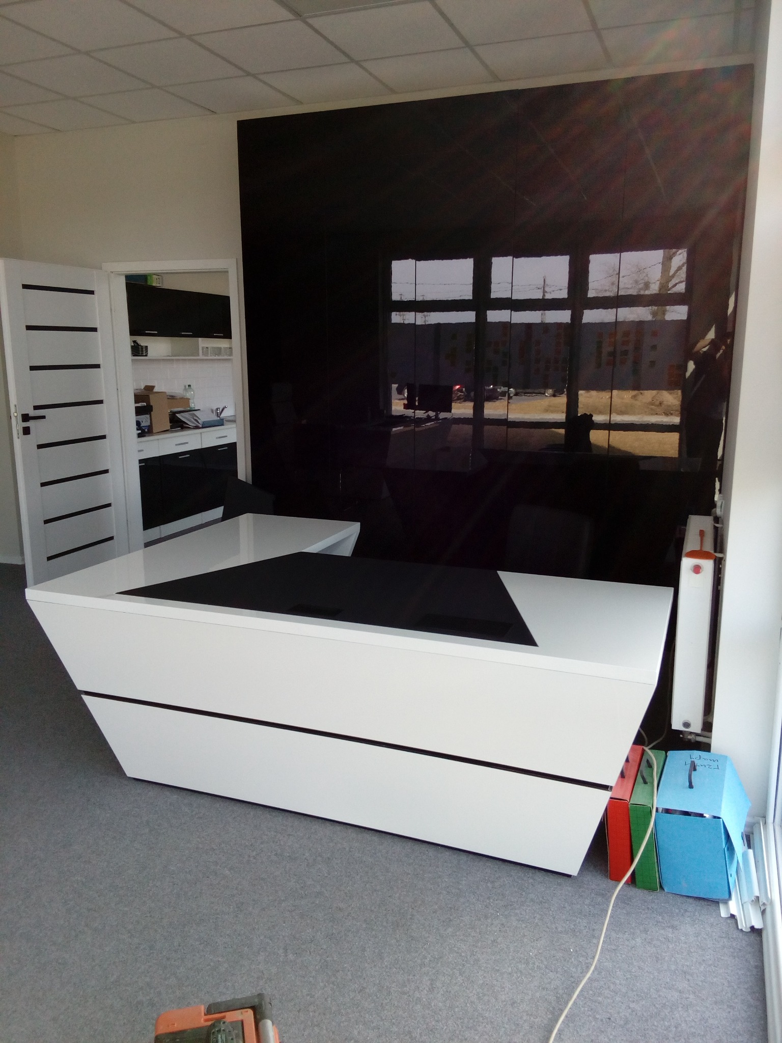 Nowoczesna, biała lada recepcyjna z czarnymi akcentami na tle czarnej, połyskującej ściany. Widoczne drzwi z czarnymi paskami i częściowo widoczna kuchnia w tle.