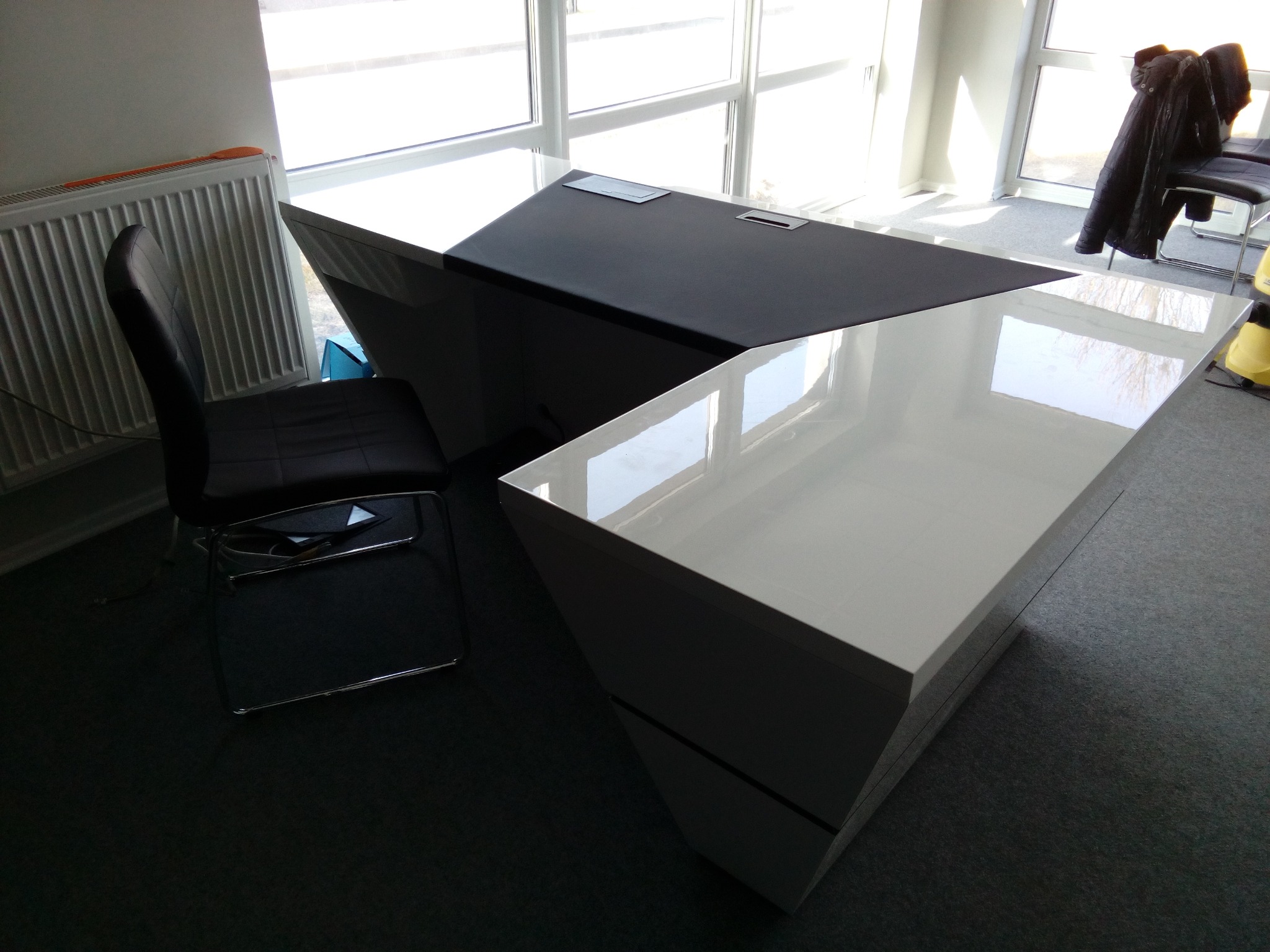 Nowoczesne biurko narożne z białym, błyszczącym blatem i czarnym elementem, uzupełnione krzesłem na chromowanym stelażu, ustawione w jasnym pomieszczeniu z oknami.