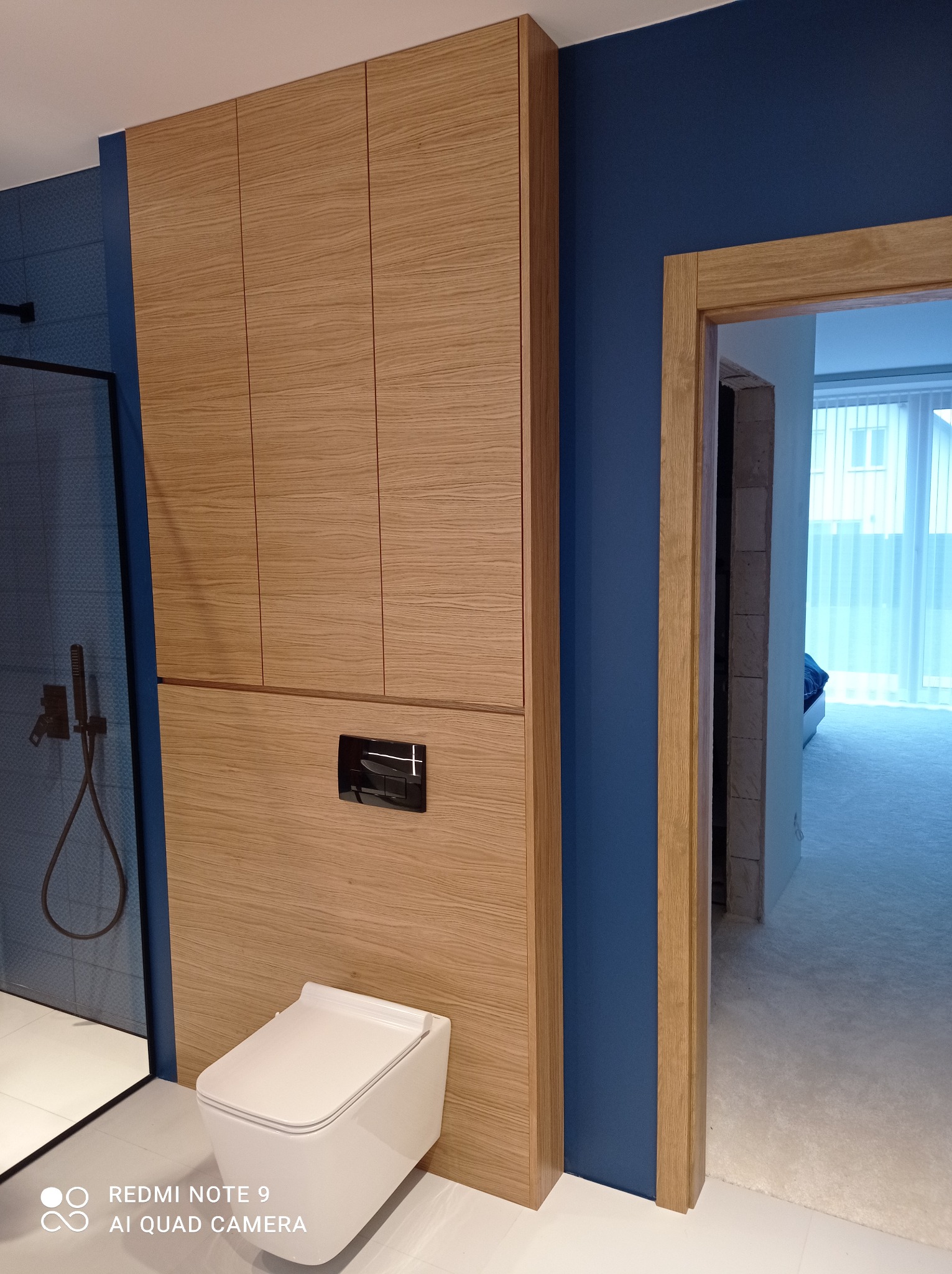 Nowoczesna łazienka z drewnianą zabudową szafy, wiszącą toaletą z czarnym przyciskiem spłukiwania i kabiną prysznicową z czarną armaturą na tle granatowej ściany i drewnianej futryny drzwi.