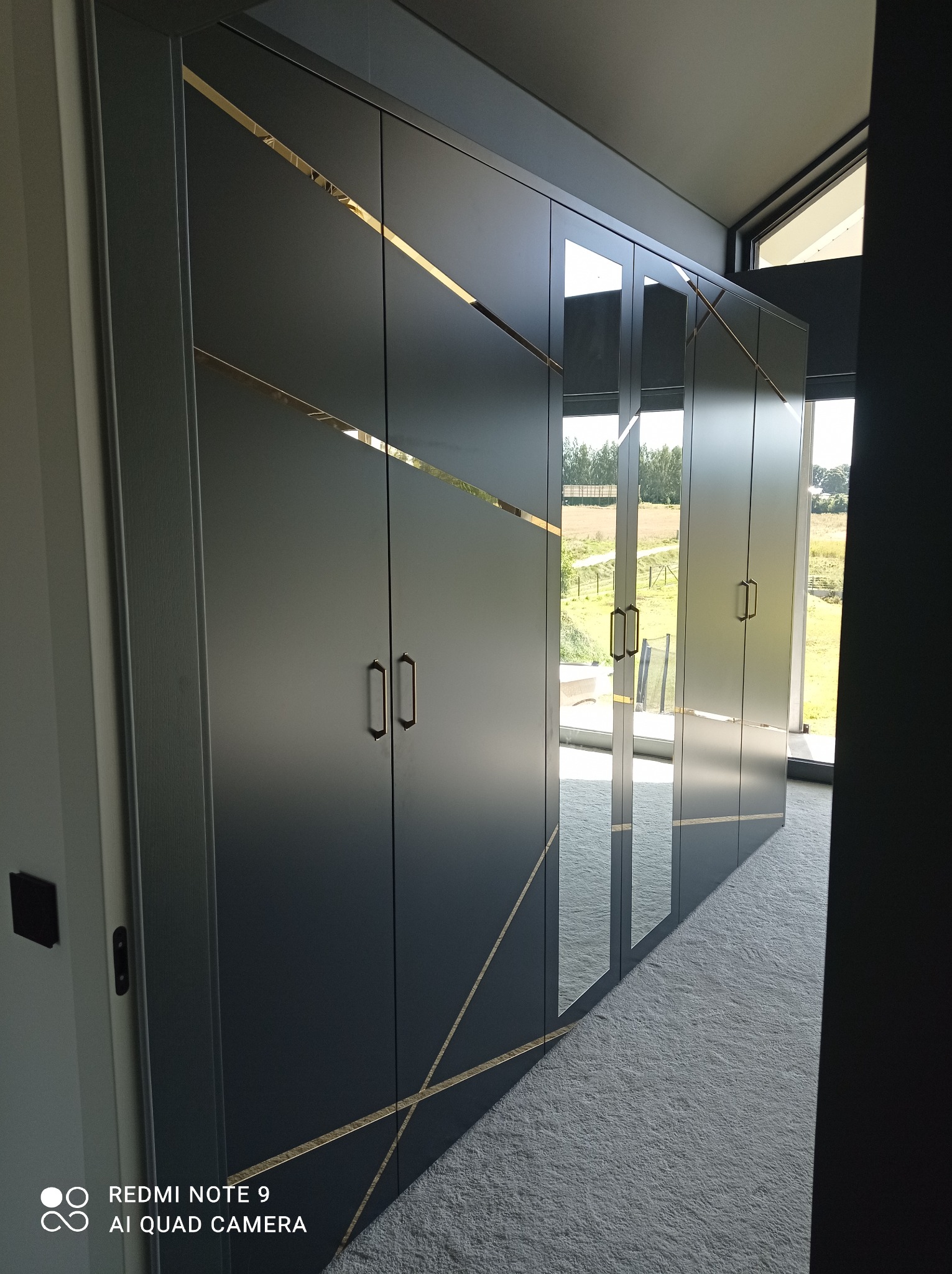 Długa, grafitowa szafa wnękowa z lustrzanymi drzwiami i złotymi akcentami, widok z boku na tle jasnego dywanu i okna z widokiem na wiejski krajobraz.