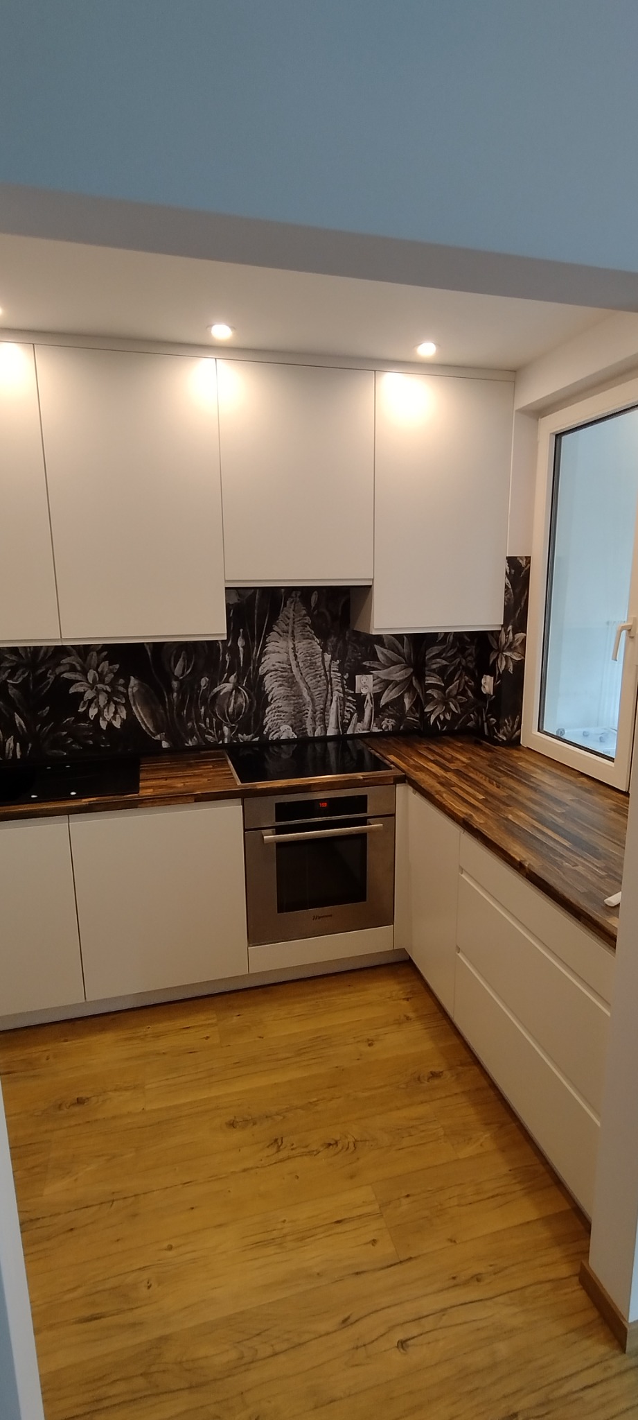 Nowoczesna kuchnia z białymi frontami szafek, drewnianym blatem i czarnym panelem ściennym w tropikalne wzory, oświetlona punktowymi lampami sufitowymi. Na blacie płyta indukcyjna i piekarnik...