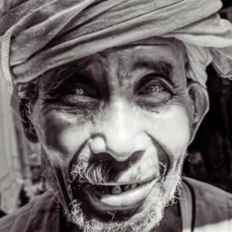 Czarno-biały portret uśmiechniętego starszego mężczyzny z turbanem, ukazujący zmarszczki, rzadki zarost i brakujące zęby, z ostrym światłem odbijającym się w jego oczach.