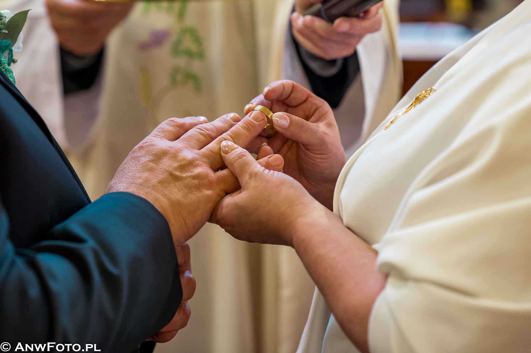 Zakładanie złotej obrączki na palec podczas ceremonii ślubnej, widoczne dłonie pary młodej i fragment stroju duchownego w tle.