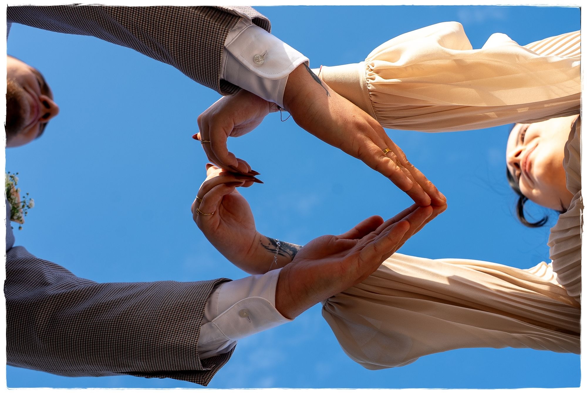Nowożeńcy tworzą serce dłońmi na tle błękitnego nieba, widok z dołu, ona w plisowanej bluzce, on w brązowej marynarce w pepitkę, widoczne obrączki i tatuaże.