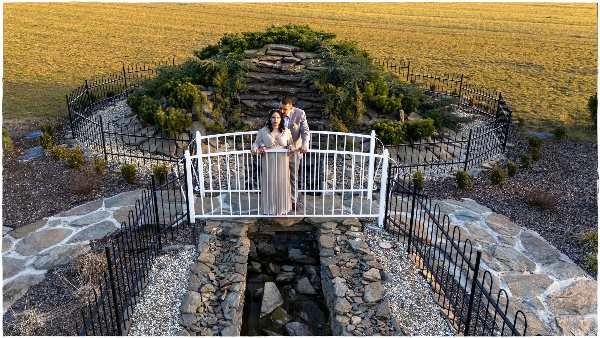 Para w eleganckich strojach stoi na białym mostku nad strumieniem otoczonym kamieniami i czarnym metalowym ogrodzeniem, w tle zielony kopiec i pole.