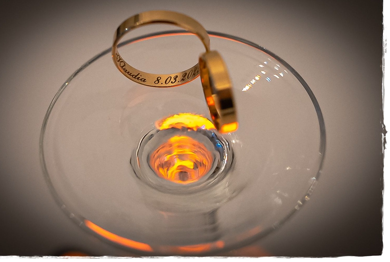 Dwa złote pierścionki z wygrawerowanym imieniem 'Klaudia' i datą '8.03.2021' leżące na krawędzi kieliszka, z refleksami światła wewnątrz.