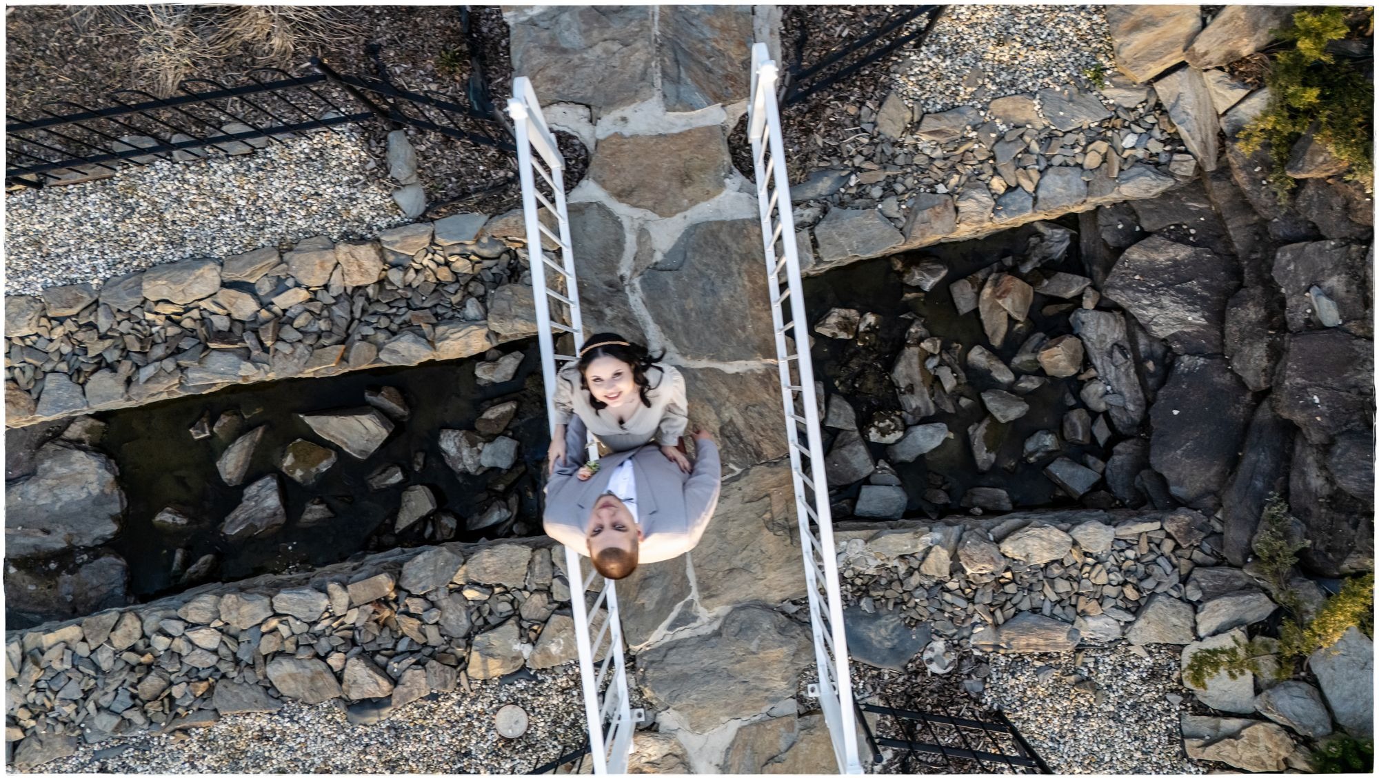 Para młoda stoi na białym mostku nad kamiennym korytem rzeki, widok z lotu ptaka, ona w jasnej sukience, on w szarym garniturze, w tle kamienne brzegi.