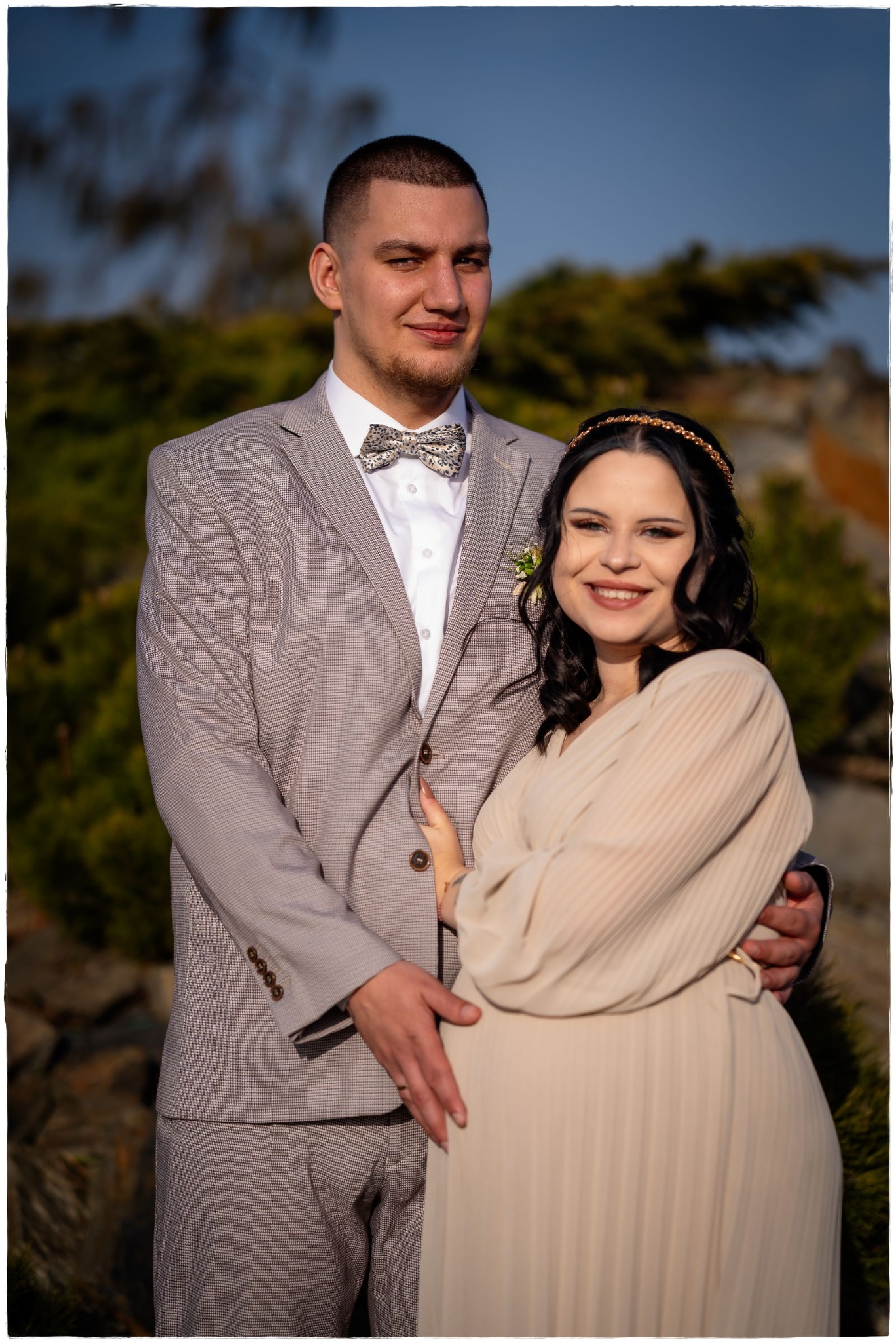 Uśmiechnięta przyszła mama w beżowej sukience i złotej opasce, obejmowana przez mężczyznę w szarym garniturze i muszce w plenerze.