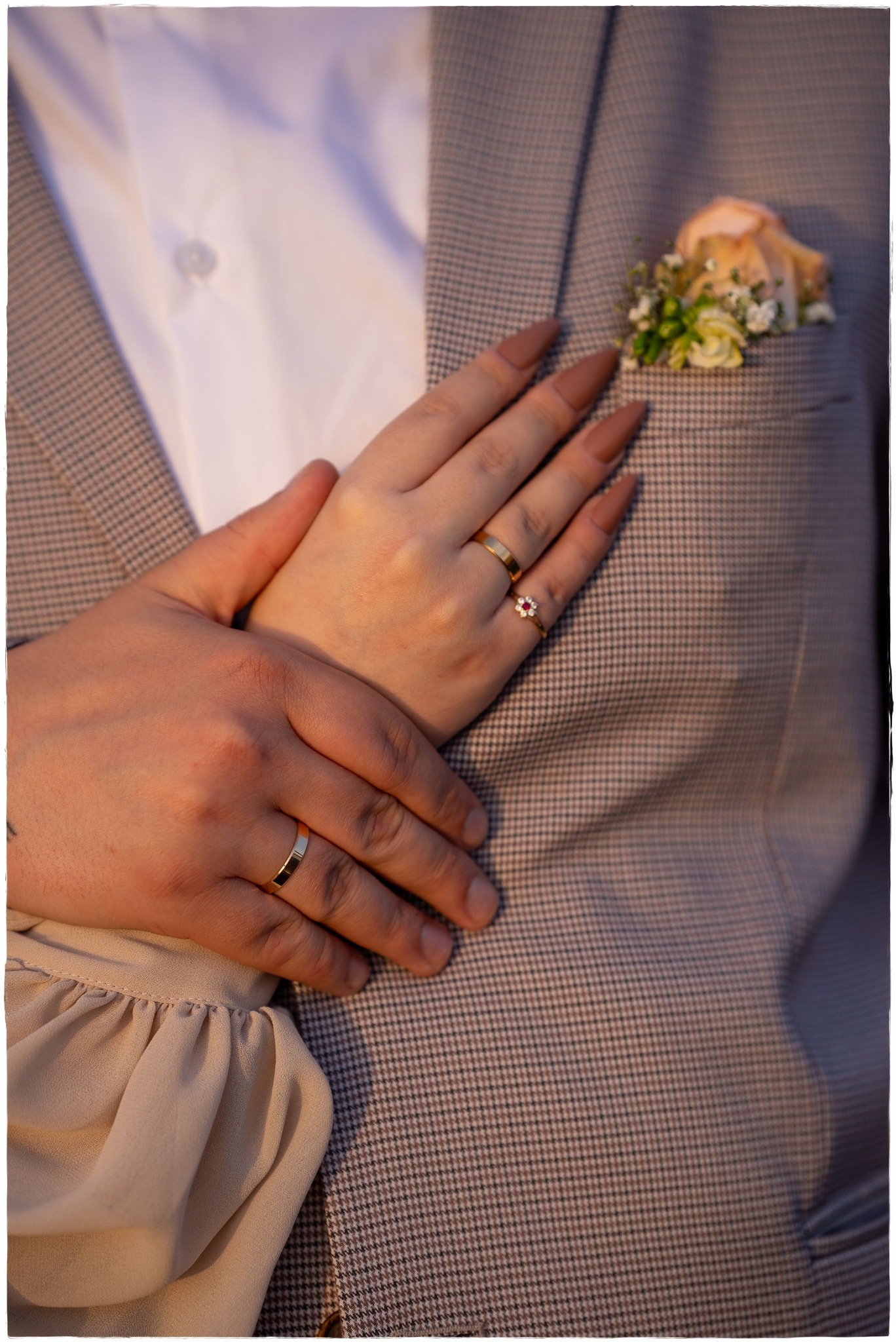 Dłonie pary z obrączkami na tle marynarki w kratę z butonierką i kremowej bluzki z bufkami.