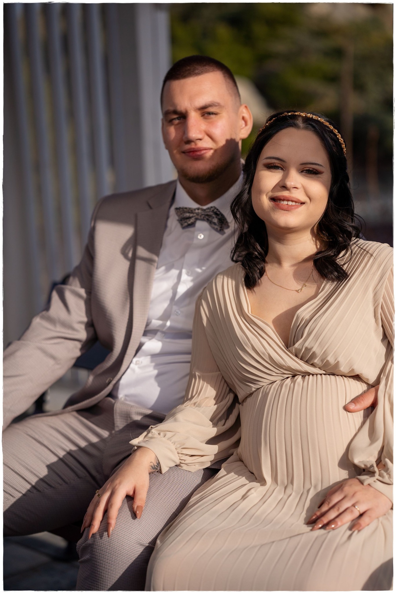 Uśmiechnięta para w plenerze: przyszła mama w beżowej sukience z plisami i złotą opaską na włosach, obok mężczyzna w szarej marynarce i muszce w kratkę.