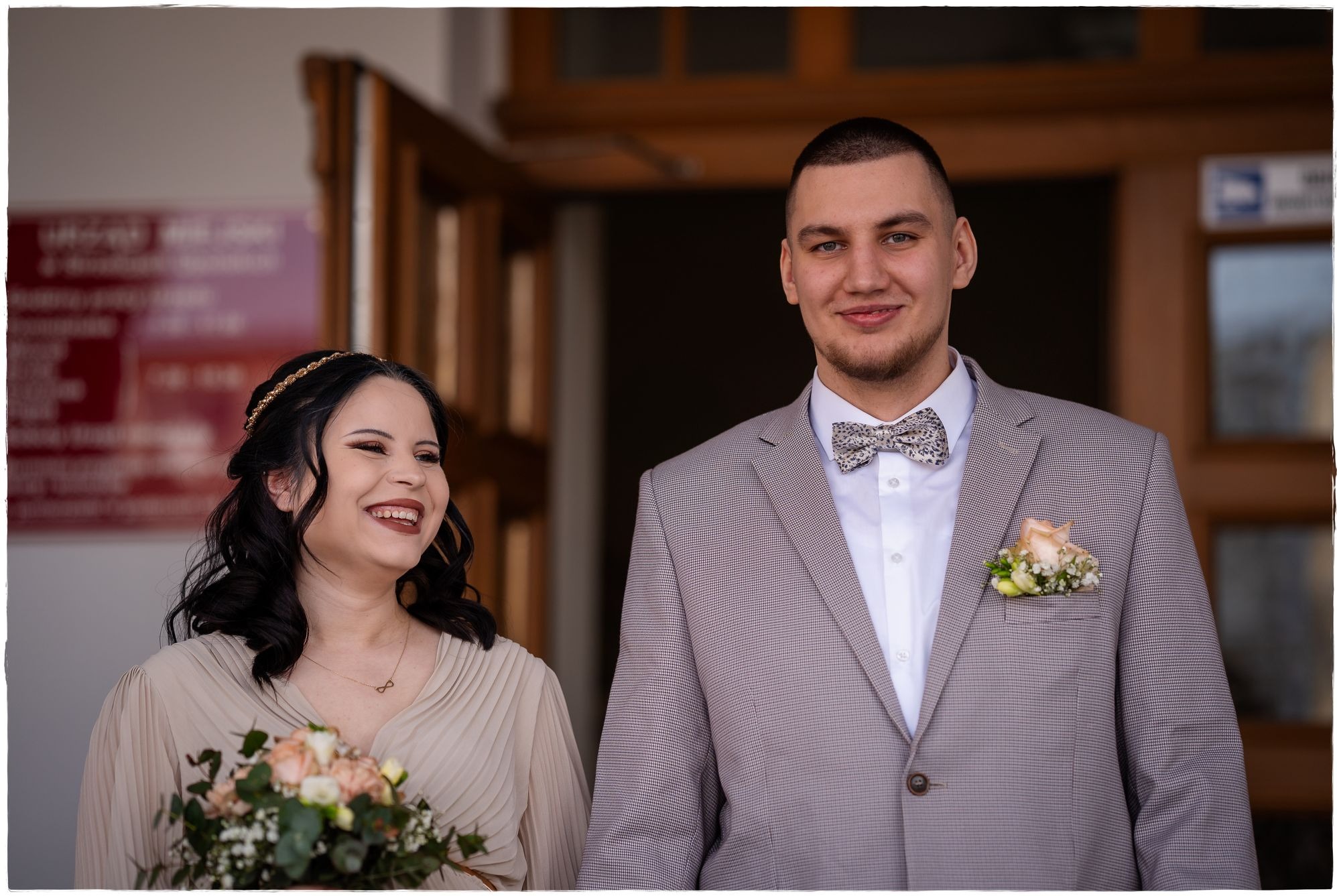 Uśmiechnięta para młoda wychodzi z budynku: panna młoda z bukietem w kremowej sukni, pan młody w szarej marynarce w drobną kratę i wzorzystej muszce.