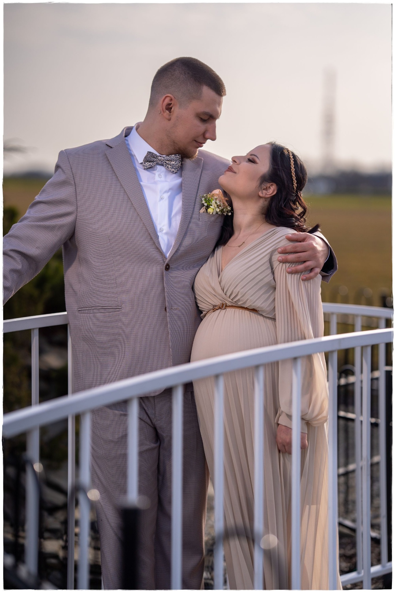 Uśmiechnięta, ciężarna kobieta w beżowej sukience, z ozdobą we włosach, opiera się na elegancko ubranym mężczyźnie w brązowym garniturze i muszce w kratkę, stoją na moście z białymi barierkami...