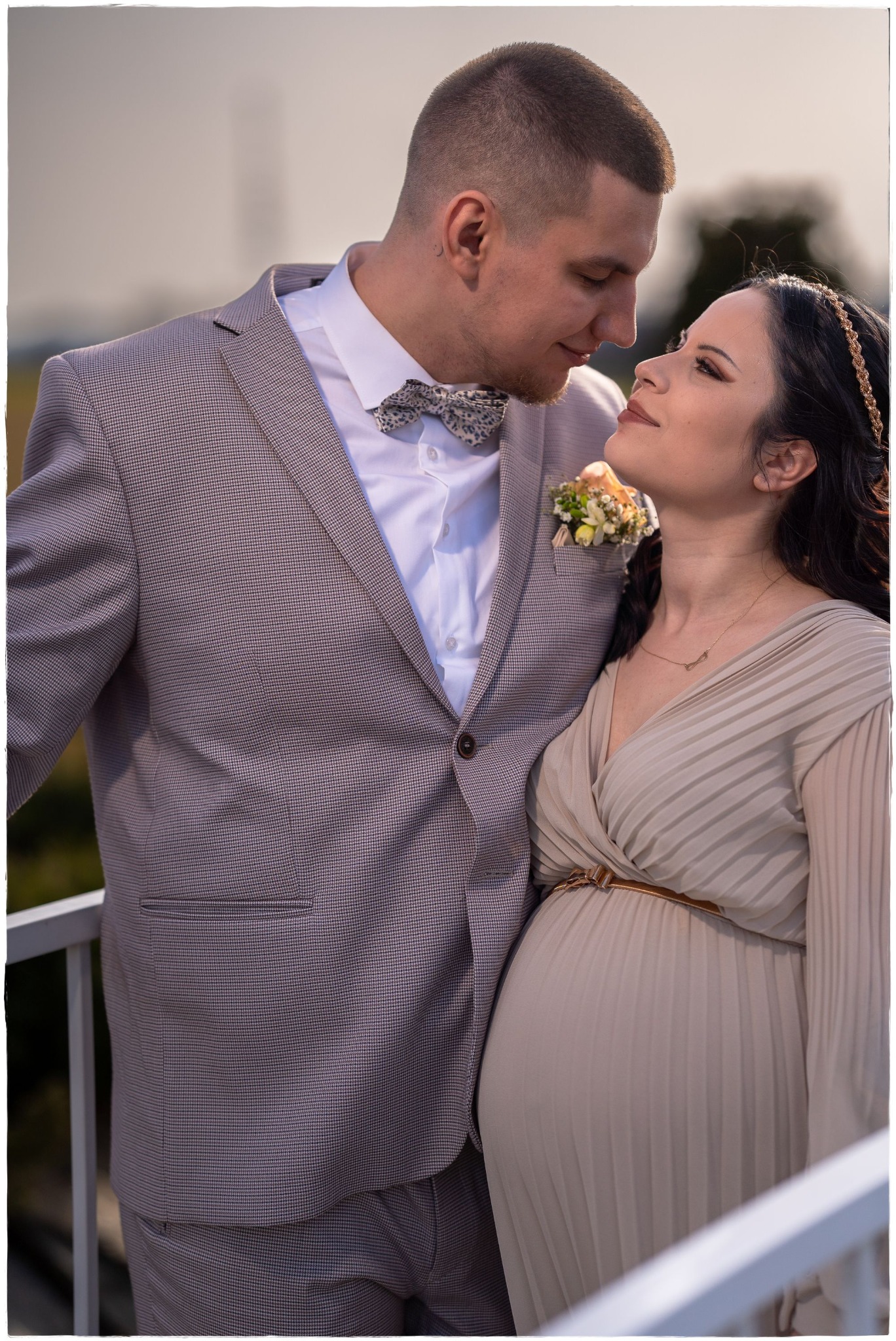 Przyszła mama w beżowej sukience, z wiankiem na włosach, patrzy z miłością na mężczyznę w szarym garniturze i muszce w kropki, stoją na balkonie.