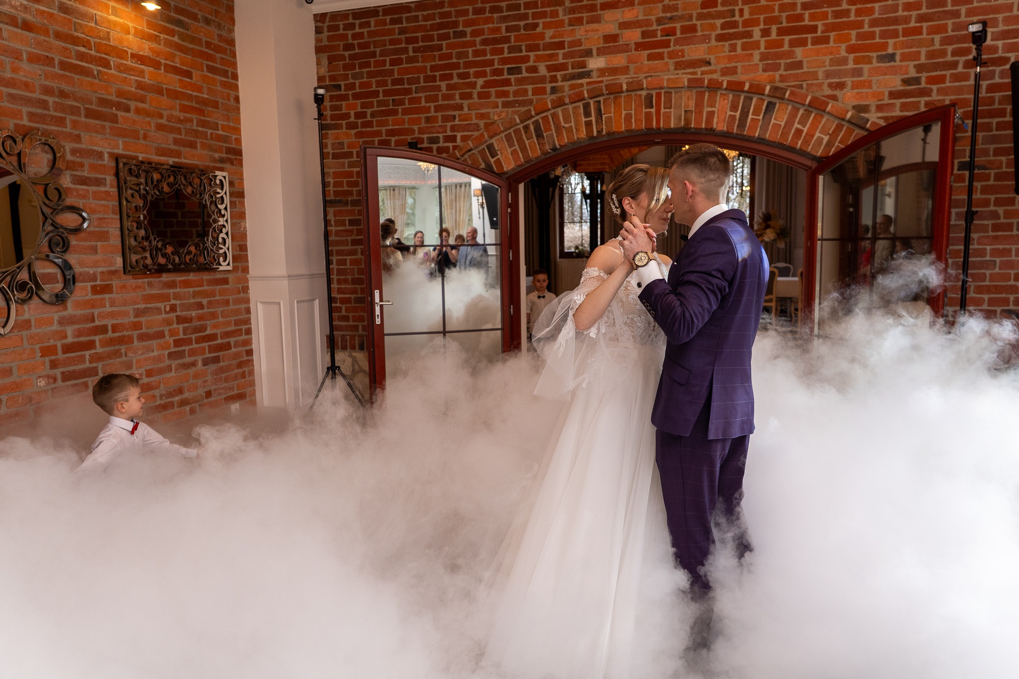 Para młoda w tańcu, otoczona sztuczną mgłą, na tle ceglanej ściany i otwartych drzwi, z widocznym chłopcem w stroju galowym.