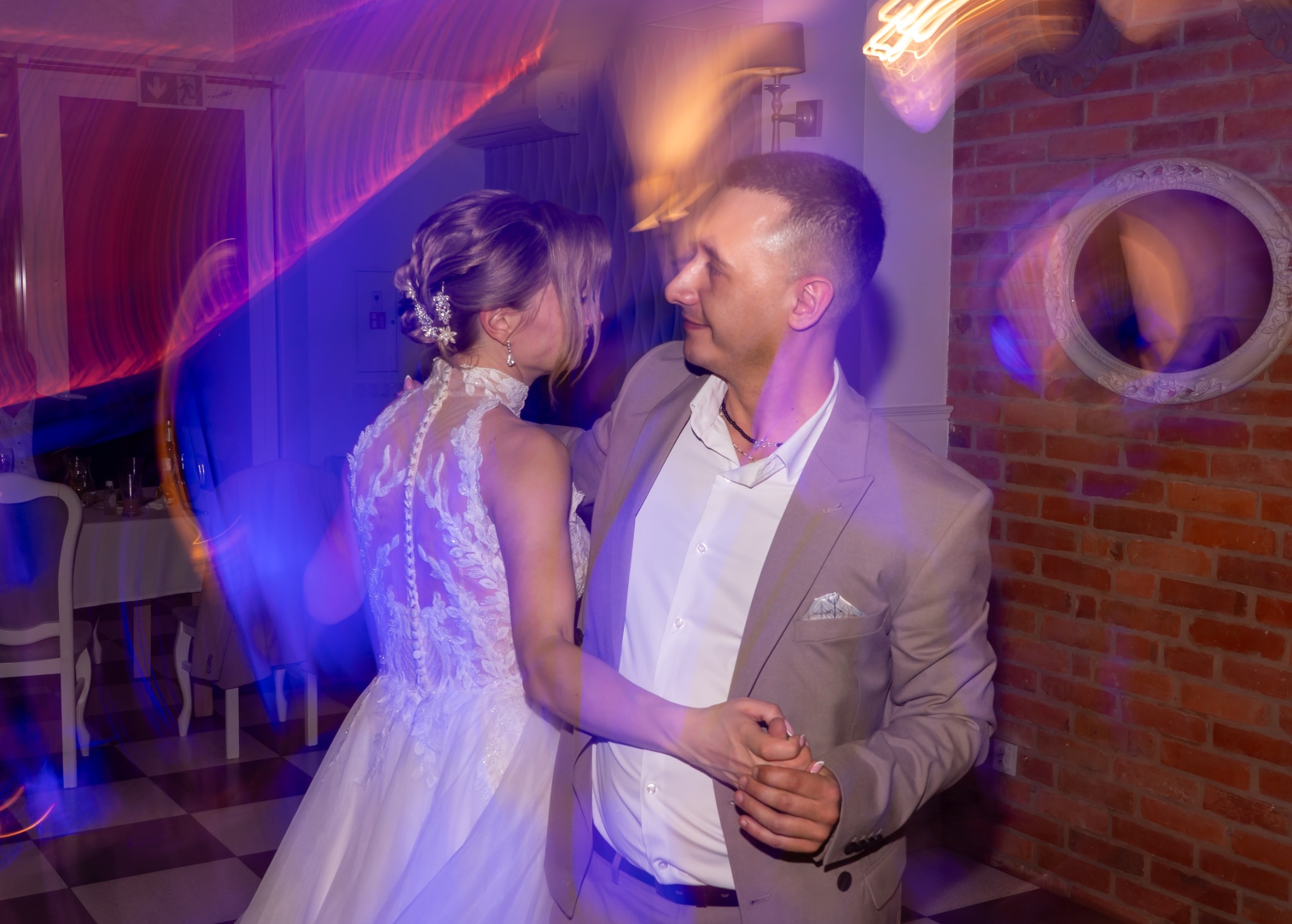 Para młoda w tańcu podczas przyjęcia weselnego, panna młoda w sukni z koronkowymi aplikacjami, pan młody w beżowym garniturze, czerwona ceglana ściana w tle, dynamiczne oświetlenie tworzy efekt...