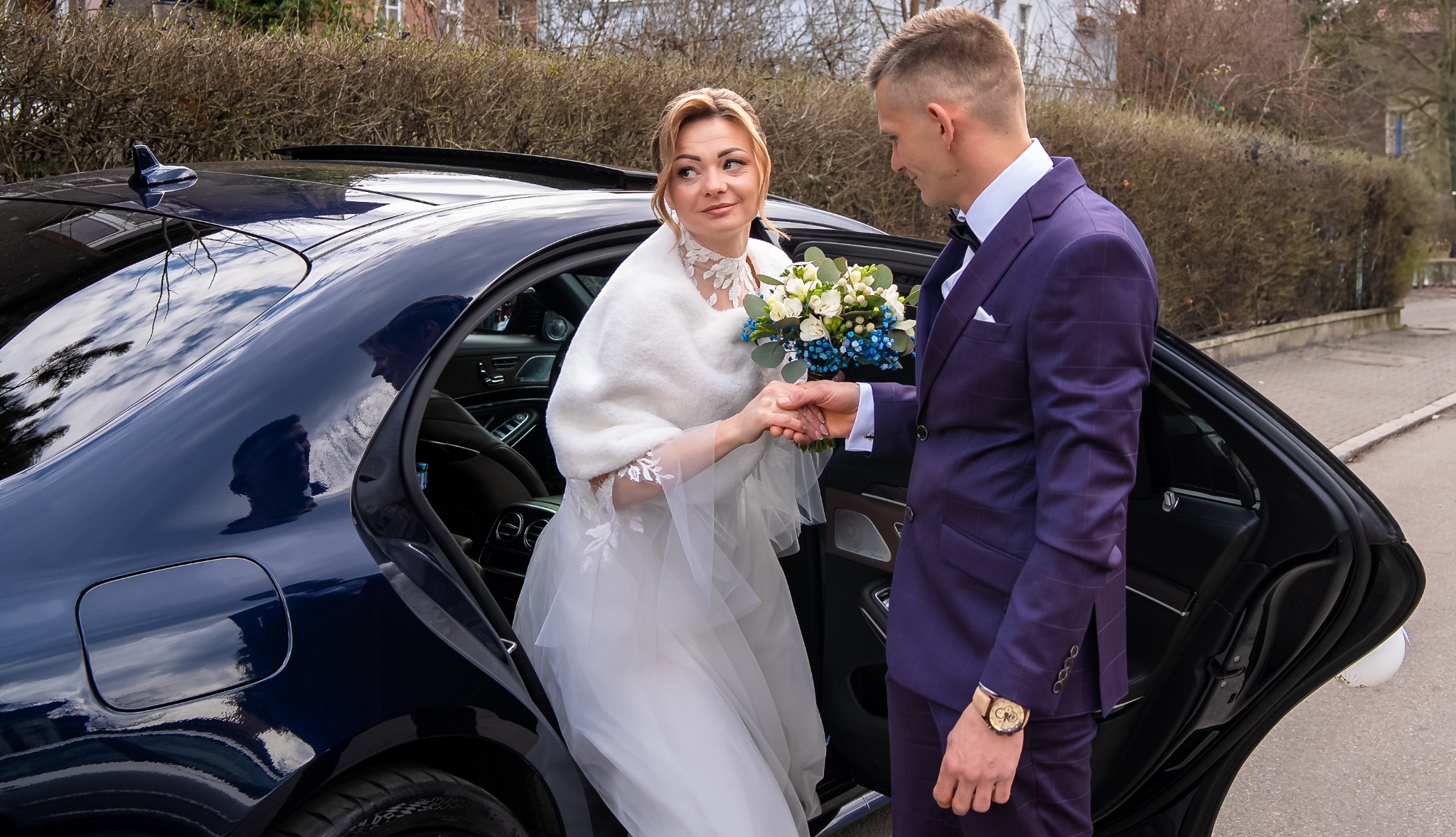 Panna młoda w białej sukni i futrzanym okryciu wychodzi z ciemnego samochodu, trzymając bukiet kwiatów, a pan młody w fioletowym garniturze podaje jej rękę.