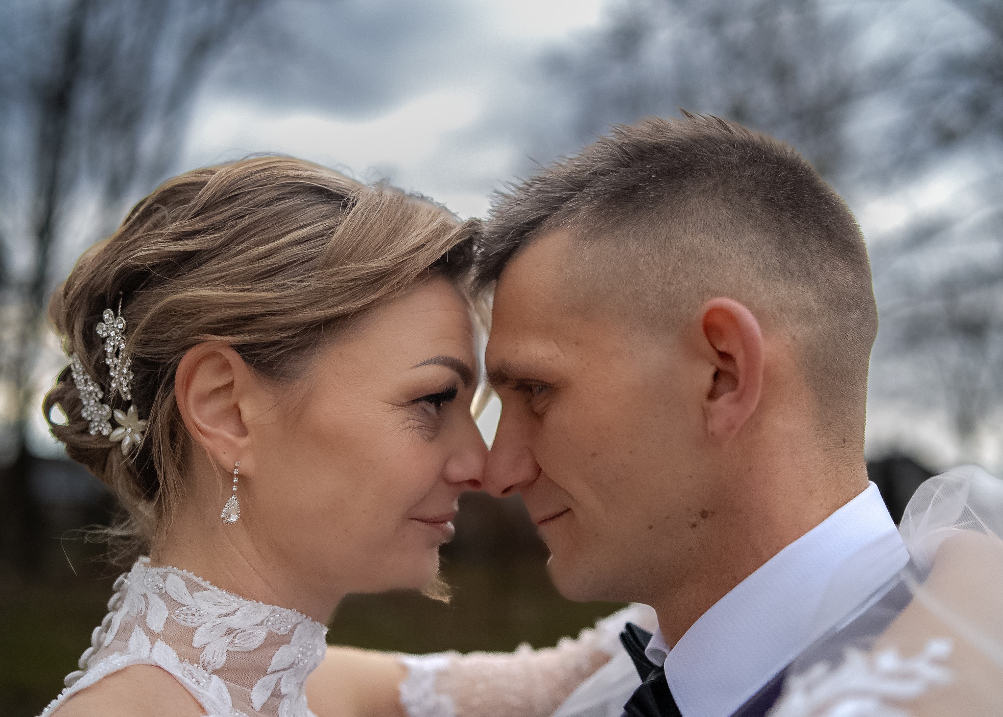 Zbliżenie na twarze pary młodej, dotykających się nosami, panna młoda z upiętymi blond włosami i ozdobną spinką, w koronkowej sukni, pan młody w czarnym smokingu i białej koszuli, tło rozmyte.