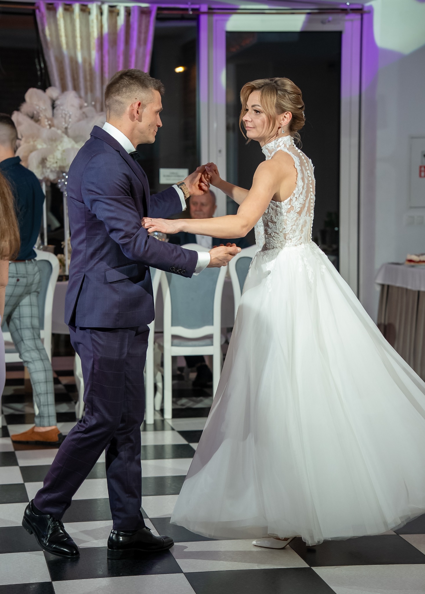 Para młoda w tańcu podczas wesela, panna młoda w sukni z koronkowym tyłem, pan młody w granatowym garniturze na szachownicowej podłodze.