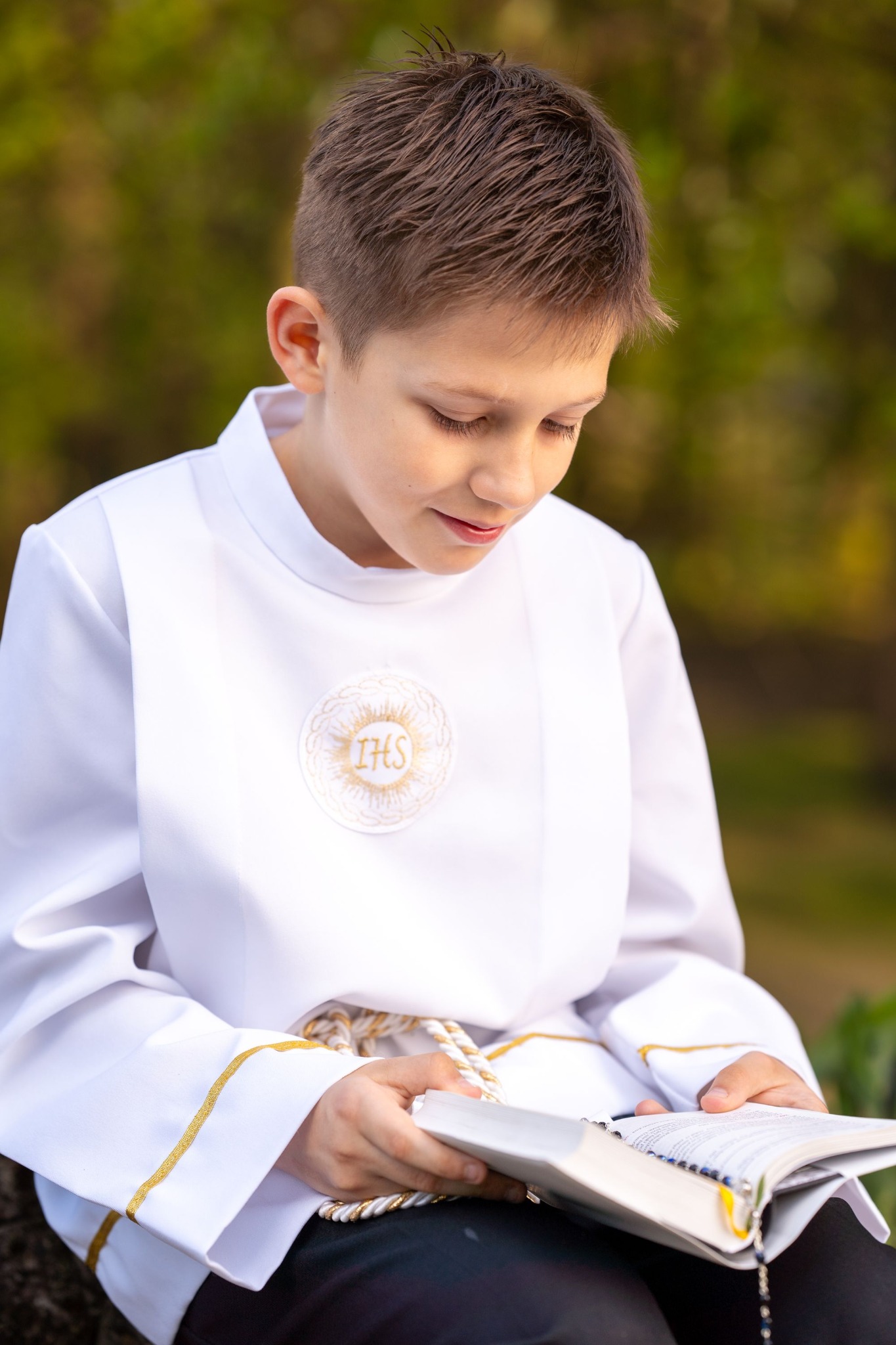 Chłopiec w białej albie z symbolem IHS czyta książkę na tle zieleni. Ujęcie z góry, miękkie światło.
