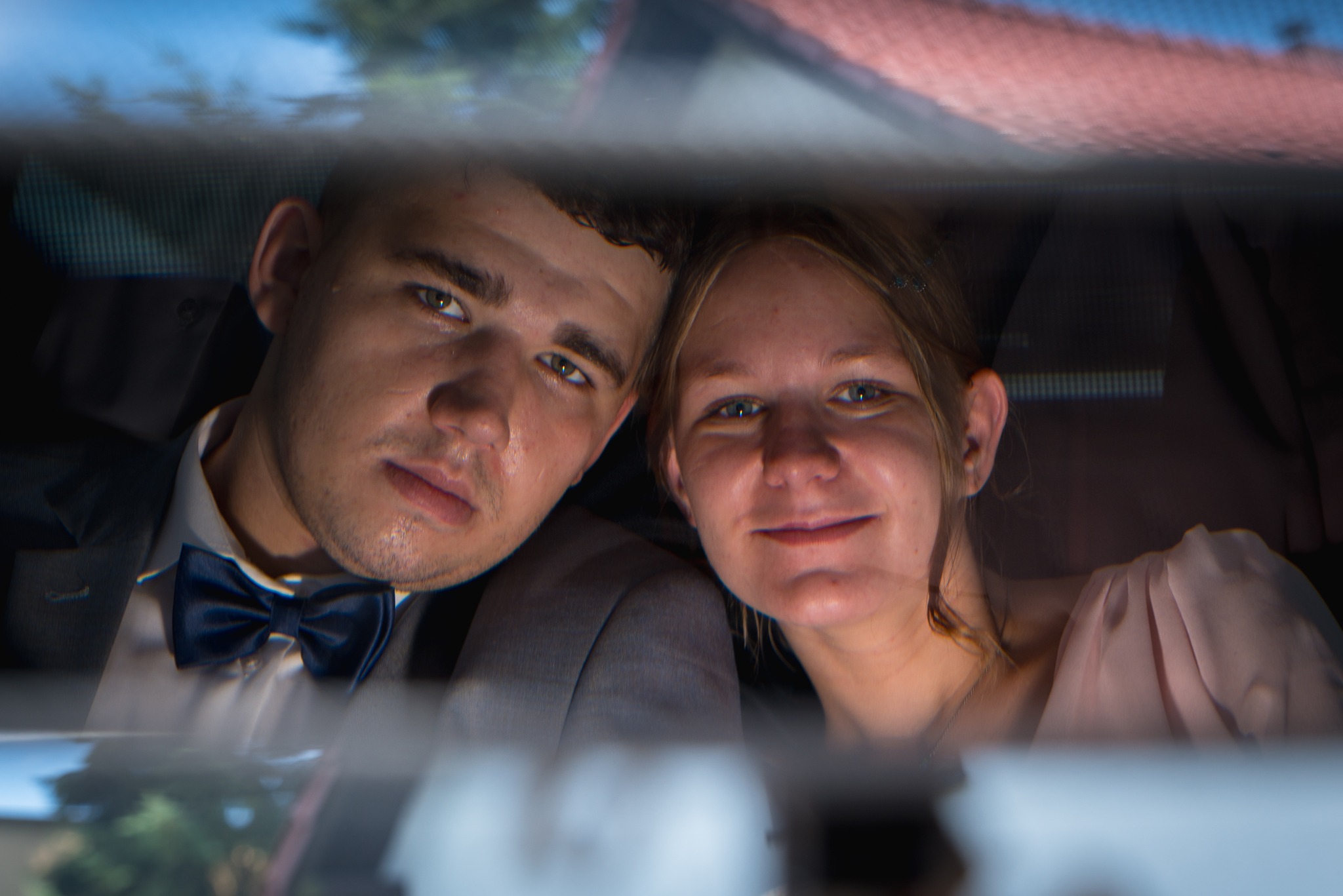 Młoda para w samochodzie, odbicie w lusterku. Mężczyzna w garniturze z muchą, kobieta w sukience. Uśmiechnięci, patrzą w obiektyw. Nieostre tło.