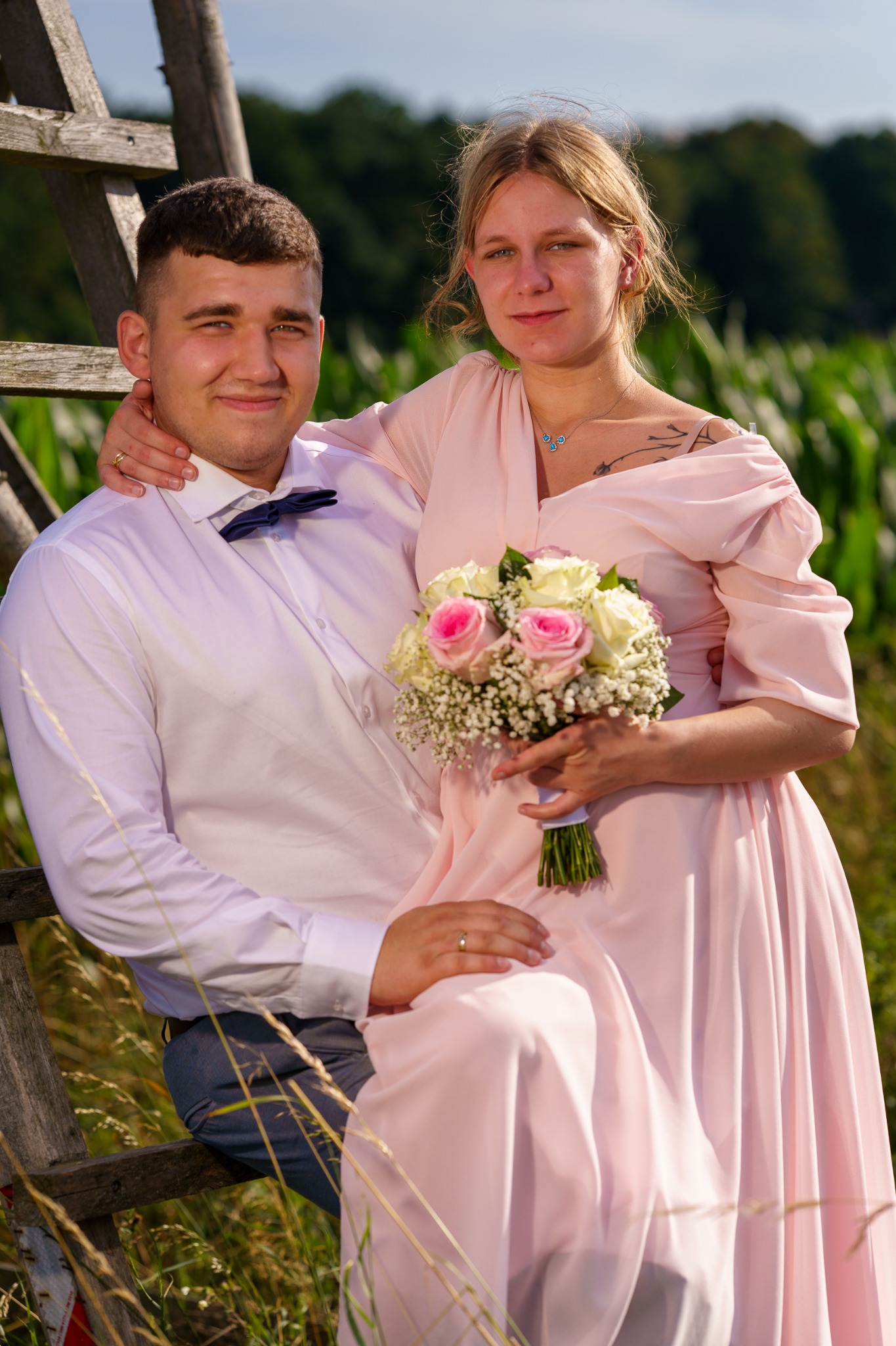 Uśmiechnięta para w pastelowych strojach pozuje z bukietem róż na tle drabiny i zieleni. Kobieta ma tatuaż na ramieniu i delikatną biżuterię.