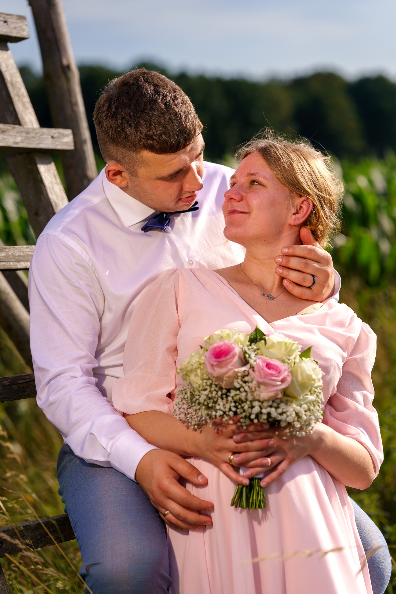 Uśmiechnięta para w objęciach, ona w pudrowej sukience z bukietem róż, on w błękitnej muszce, opierają się o drewnianą konstrukcję na tle zieleni.