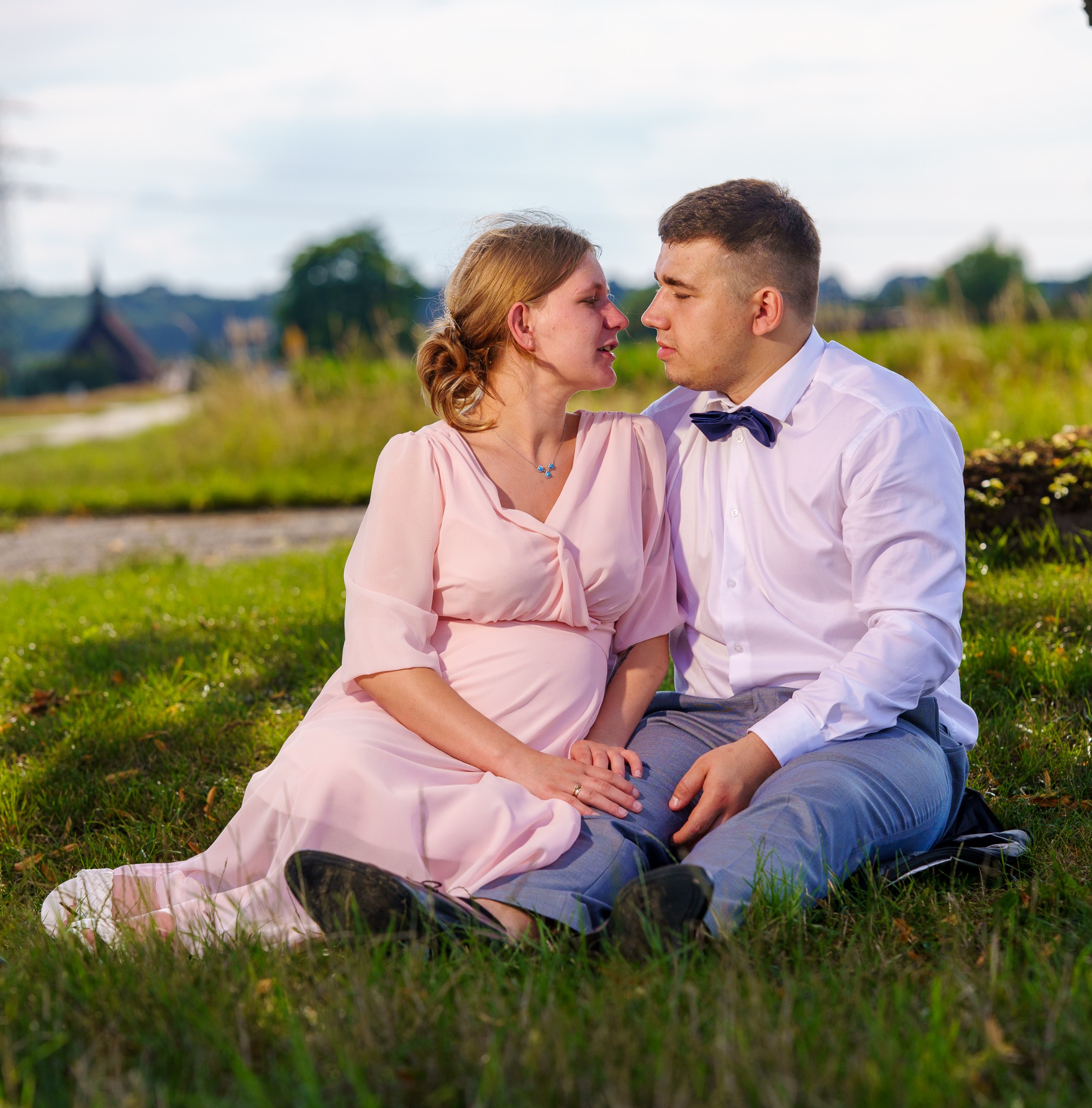Para siedzi na trawie, kobieta w różowej sukience, mężczyzna w koszuli i muszce, patrzą na siebie. Sesja w plenerze, w tle zieleń i zabudowania.