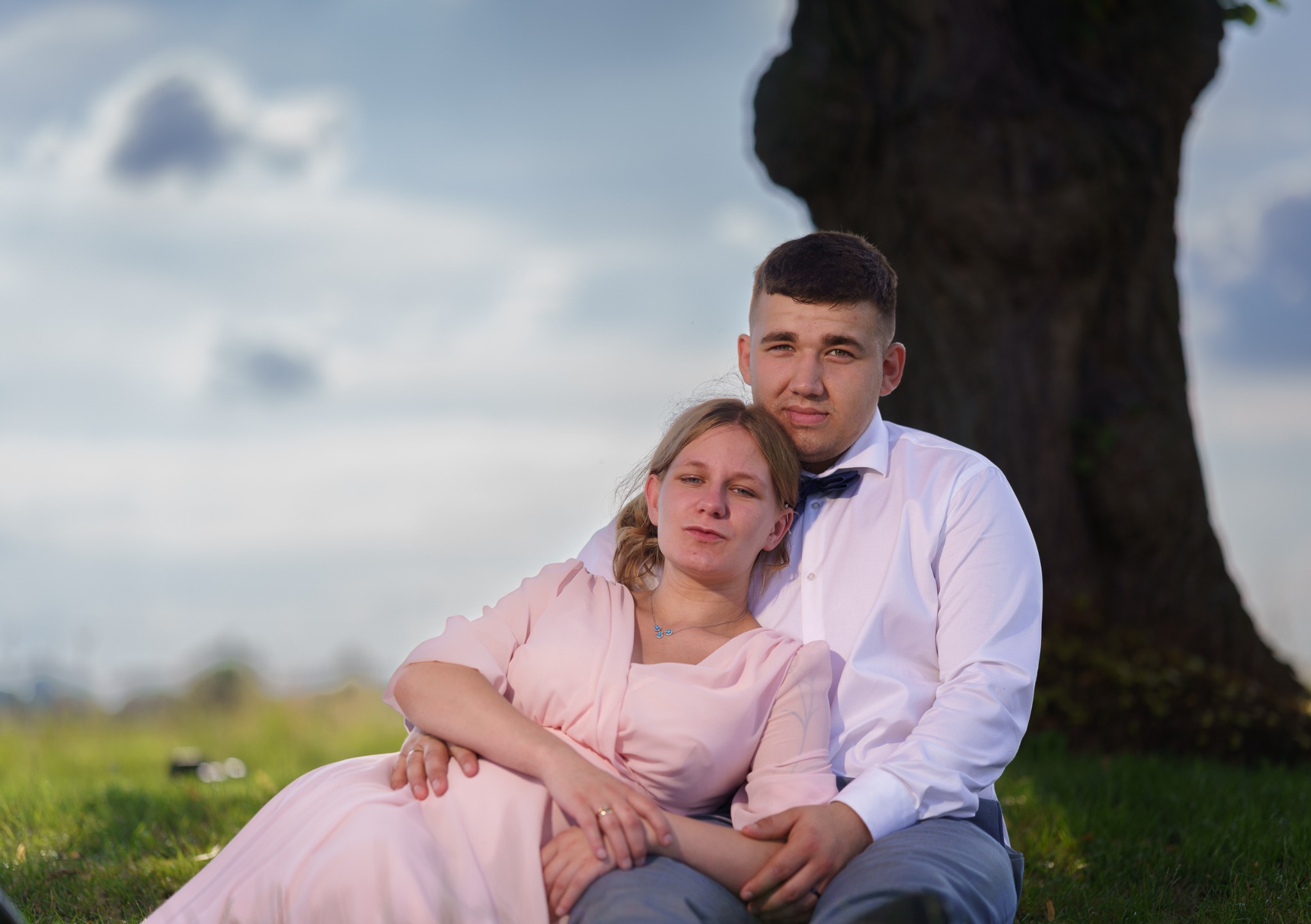 Para siedzi na trawie pod drzewem. Kobieta w pudrowo różowej sukience, mężczyzna w białej koszuli i granatowej muszce. Romantyczna sesja plenerowa z naturalnym światłem.