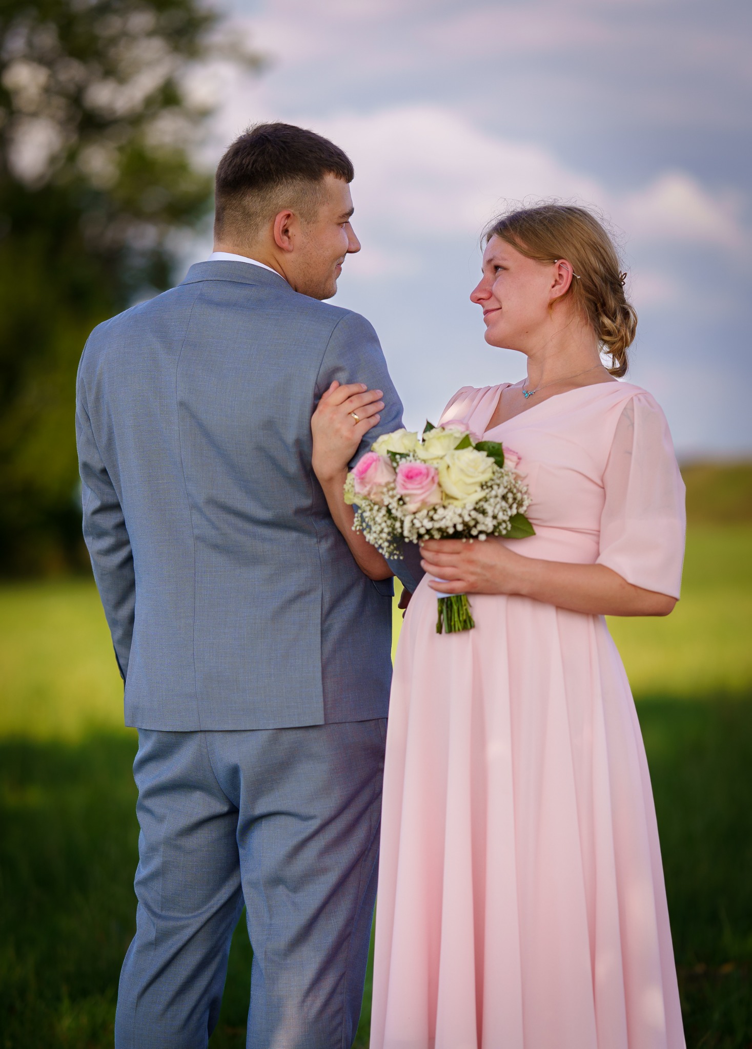 Para młoda w plenerze: on w szarym garniturze, ona w różowej sukience trzyma bukiet róż. Uśmiechnięci patrzą na siebie, w tle zieleń drzew i błękitne niebo.