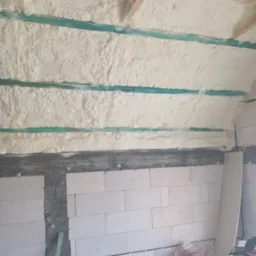 Ściana i sufit w trakcie prac izolacyjnych, widoczna warstwa pianki między drewnianymi belkami oraz fragment ściany z białych bloczków.