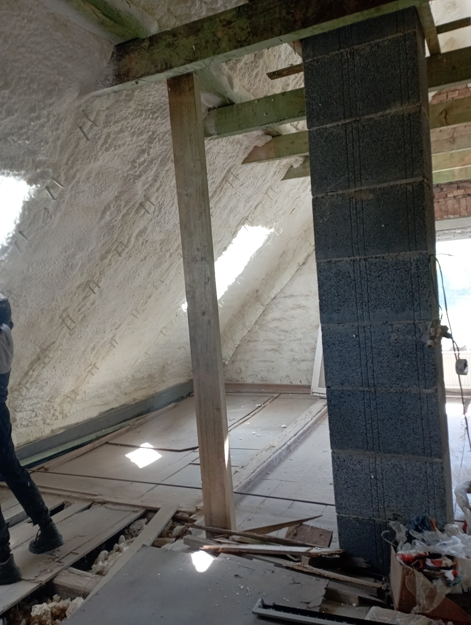 Poddasze w trakcie ocieplania, widoczne krokwie, drewniany słup, murowany filar z bloczków i biała pianka izolacyjna na skośnych ścianach.