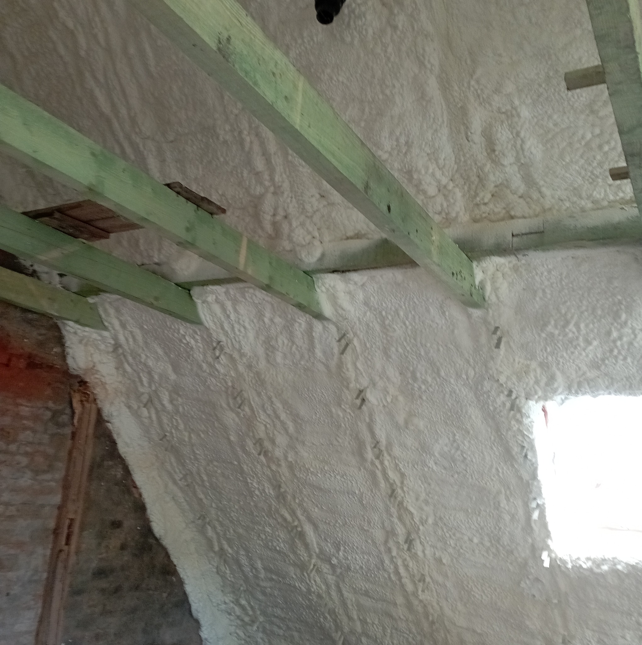 Ściana i poddasze pokryte grubą warstwą białej izolacji natryskowej, widoczne zielone drewniane belki konstrukcyjne i fragment okna.