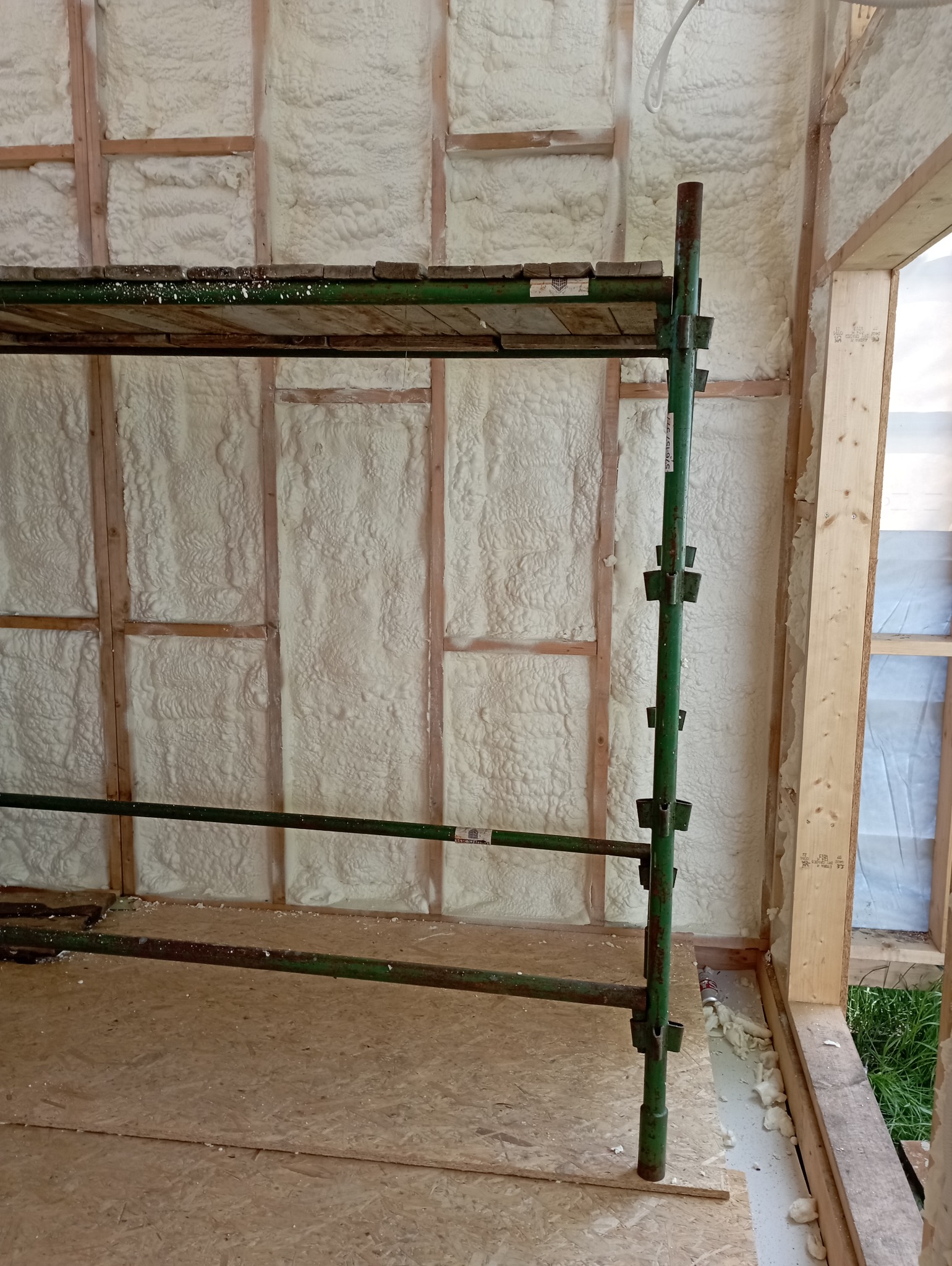 Ściana wewnętrzna budynku z drewnianą konstrukcją szkieletową wypełniona białą pianą izolacyjną; widoczne zielone rusztowanie i podłoga z płyt OSB.