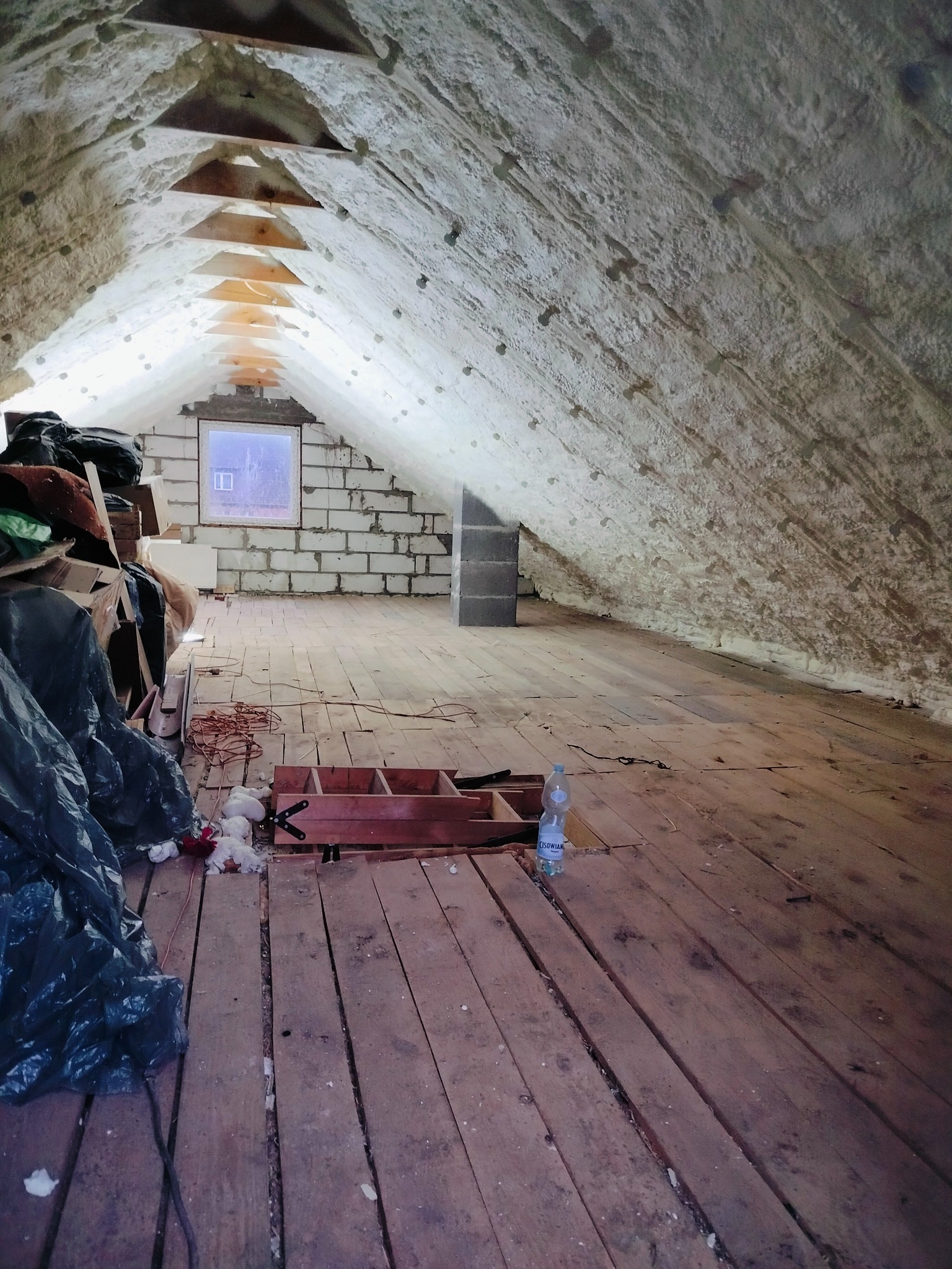 Poddasze po natrysku izolacji, drewniana podłoga z widocznymi szczelinami, okno w ścianie z pustaków, część poddasza zagracona workami i przedmiotami.