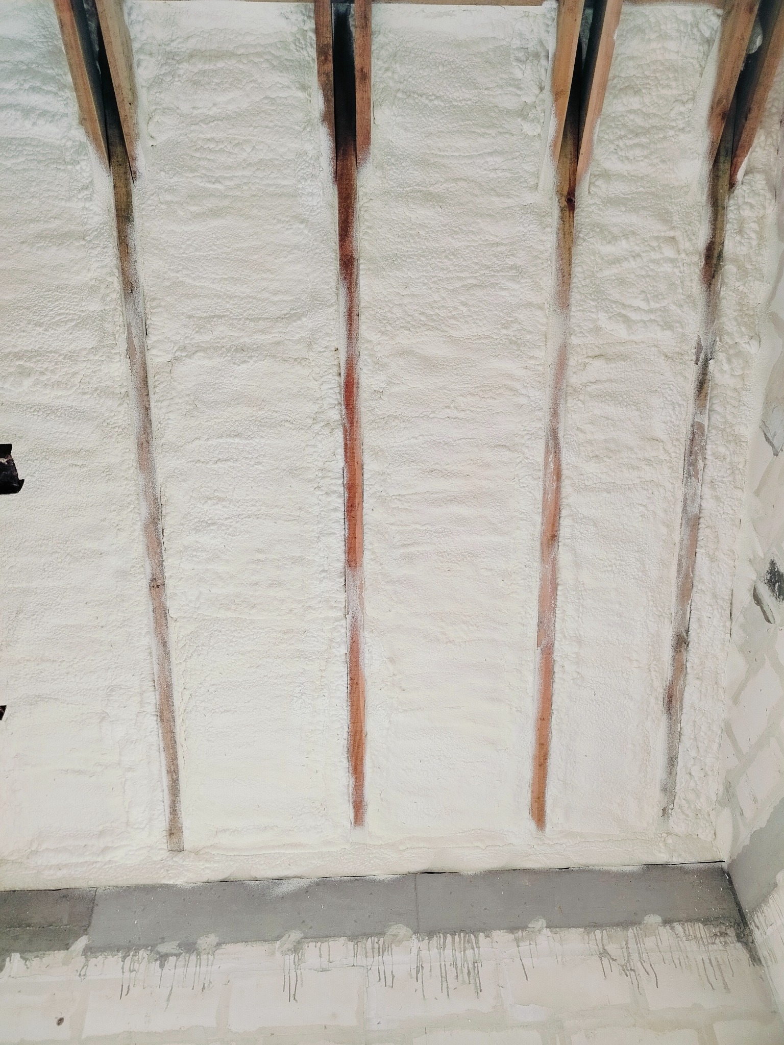 Ocieplony pianką strop z drewnianymi belkami, widoczne nierówności struktury piany i ślady po montażu.