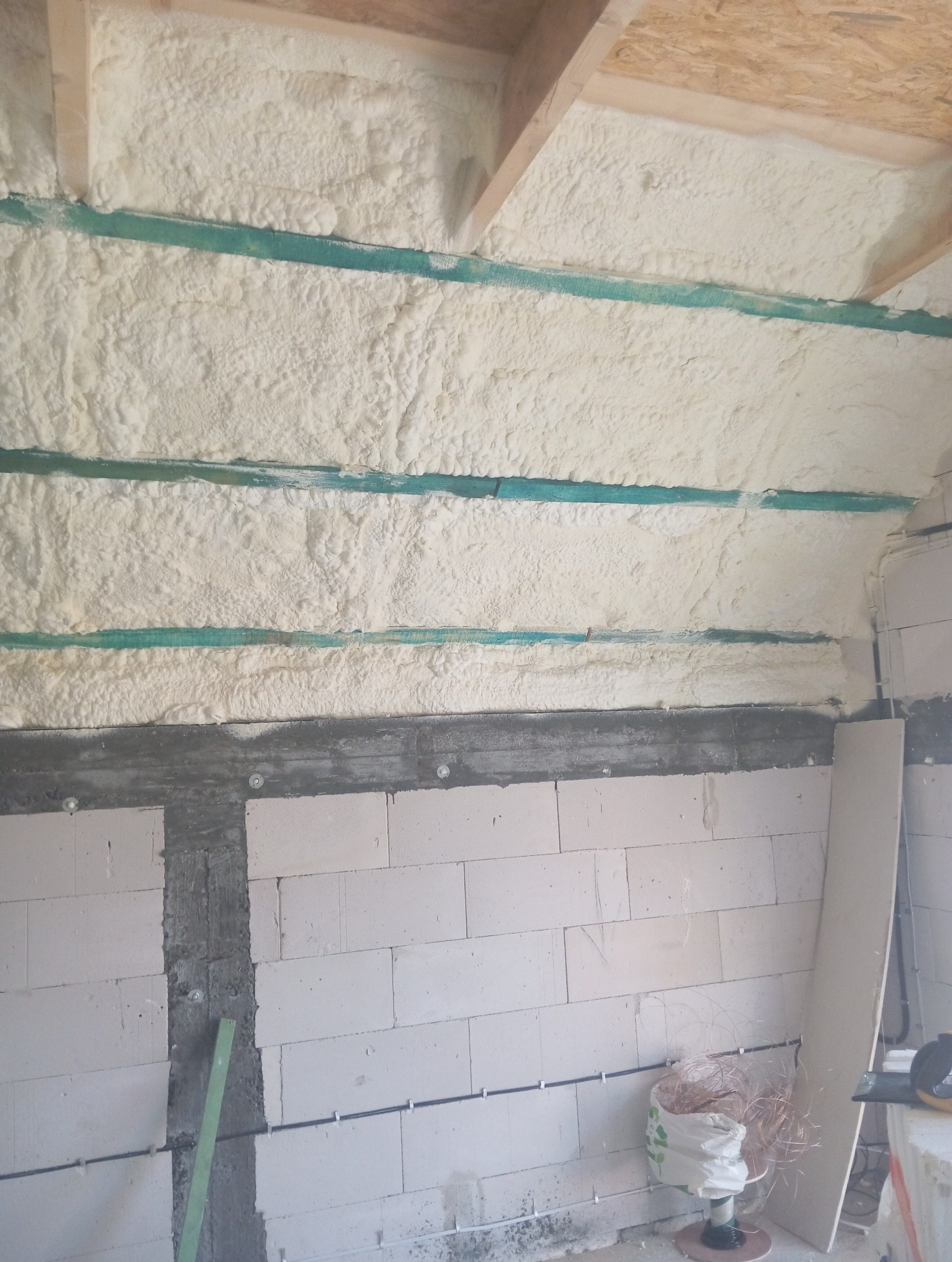 Ściana i sufit w trakcie prac izolacyjnych, widoczna warstwa pianki między drewnianymi belkami oraz fragment ściany z białych bloczków.