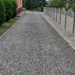 Długa, szara droga z kruszywa prowadząca do domu, obok ozdobny betonowy płot i zadbane tuje.