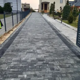 Długa, symetryczna aleja z szarej kostki brukowej prowadząca do domu, obrzeża z ciemniejszej kostki, ogrodzenie po lewej stronie, drzewa i krzewy po obu stronach.