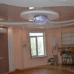 Jasny pokój z drewnianą podłogą, błyszczącym sufitem podwieszanym z ozdobnym żyrandolem, klimatyzacją na ścianie i niedokończoną zabudową kuchenną w tle.