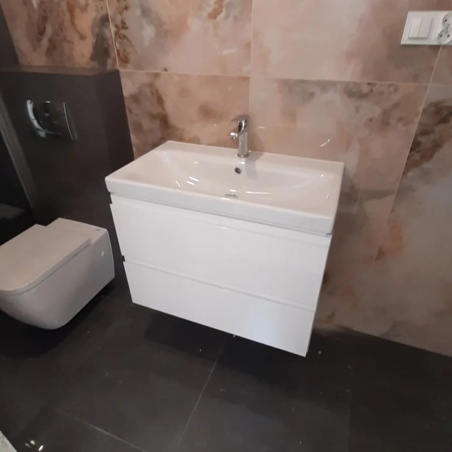 Nowoczesna łazienka z białą szafką pod umywalkę i wiszącą toaletą, wykończona płytkami imitującymi marmur w odcieniach beżu i brązu.