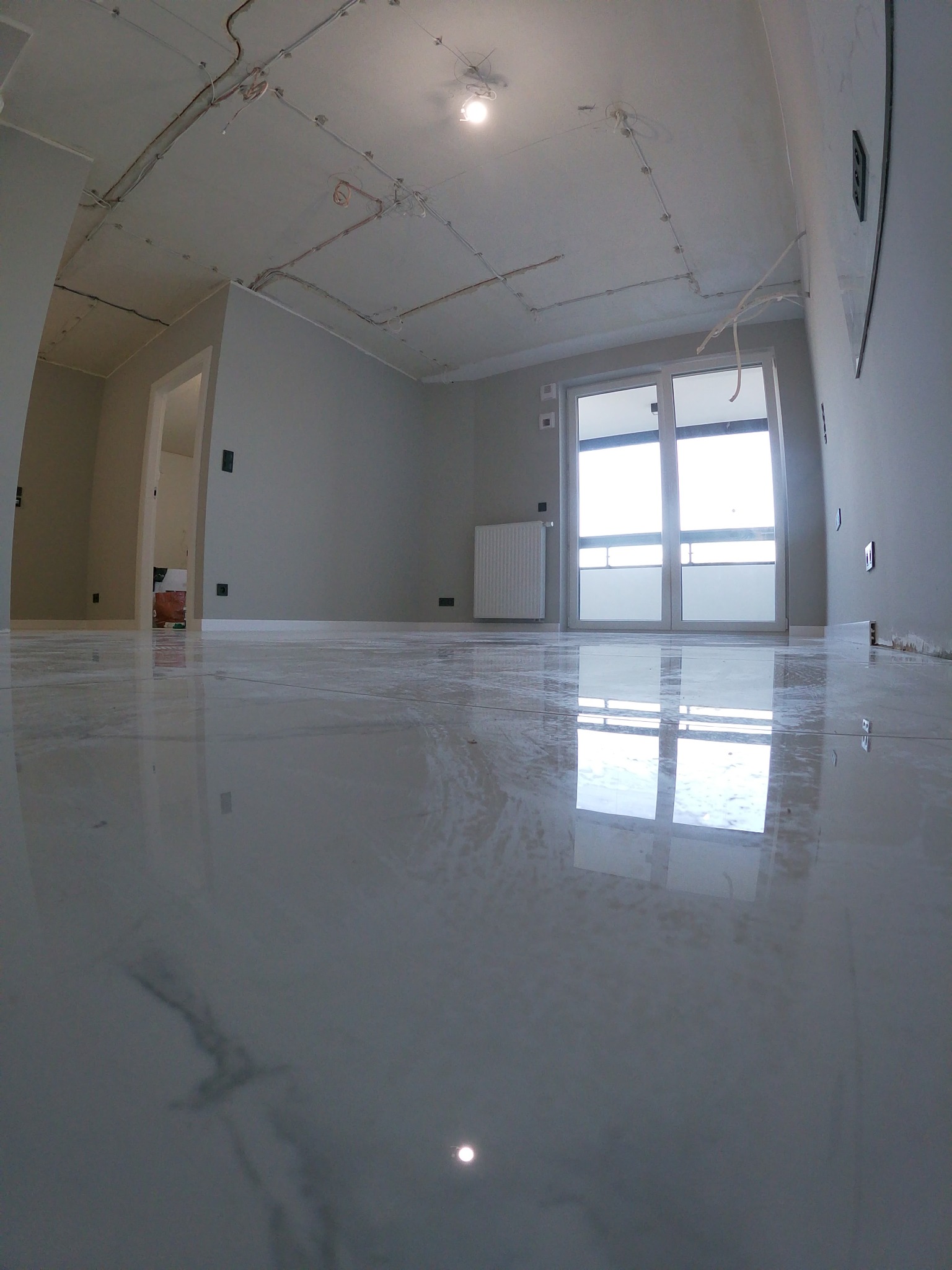 Wnętrze w trakcie prac wykończeniowych: położona, błyszcząca podłoga z wzorem marmuru, ściany w odcieniach szarości, widoczne instalacje elektryczne na suficie, grzejnik pod oknem balkonowym.