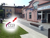 Budynek z szyldem 'Optyk' i biura nieruchomości DGR, widok z zewnątrz, chodnik z ławkami, trawnik, drzewa, w tle zabudowa miejska.