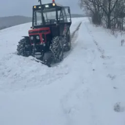 Czerwony traktor Zetor 7745 z pługiem odśnieżającym toruje drogę przez zasypane śniegiem pole, w tle zalesione wzgórza i budynek.